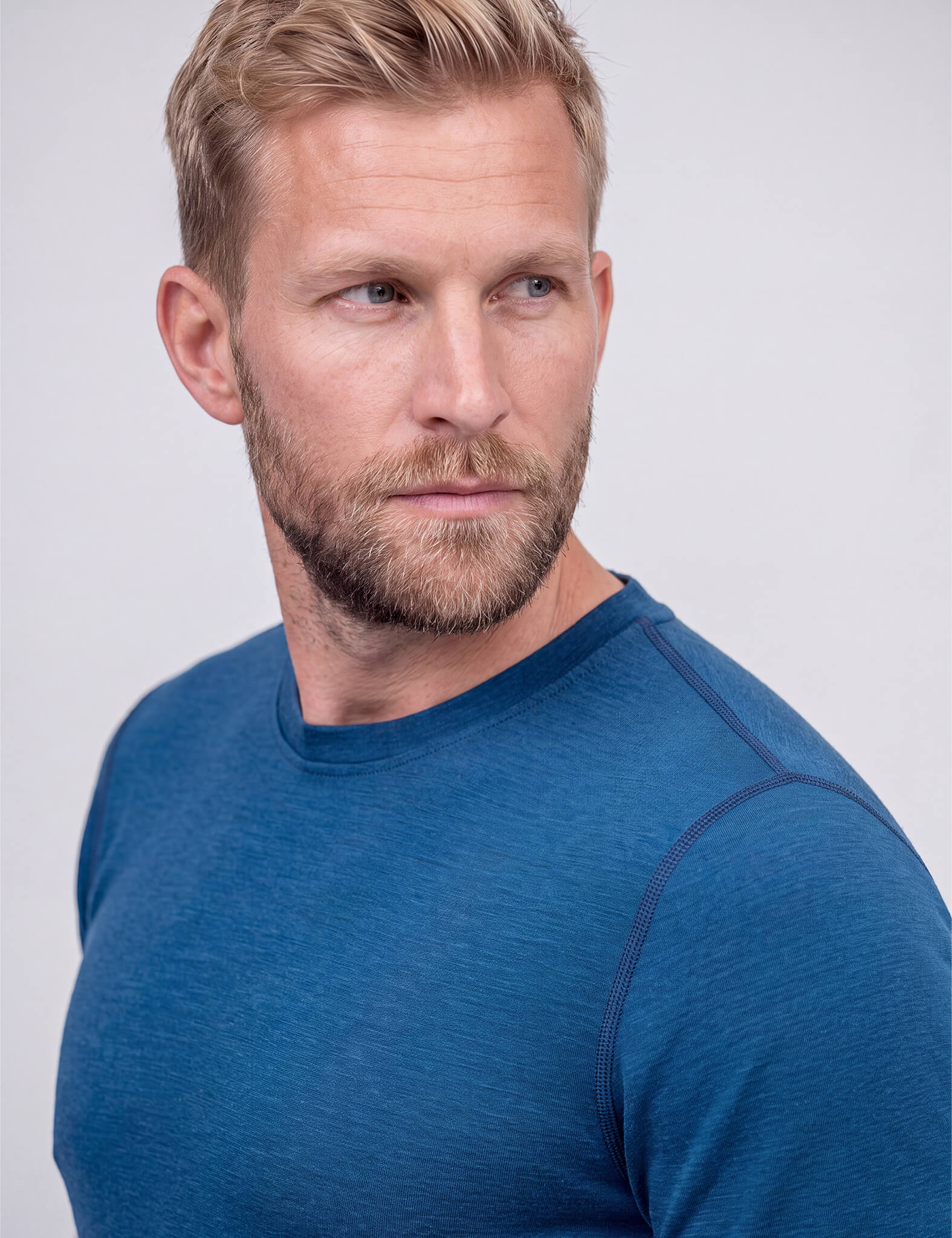 A blond man with a beard, wearing the Crew Neck – Classic Merino 160 Men in blue, looks to his left against a plain light gray background.