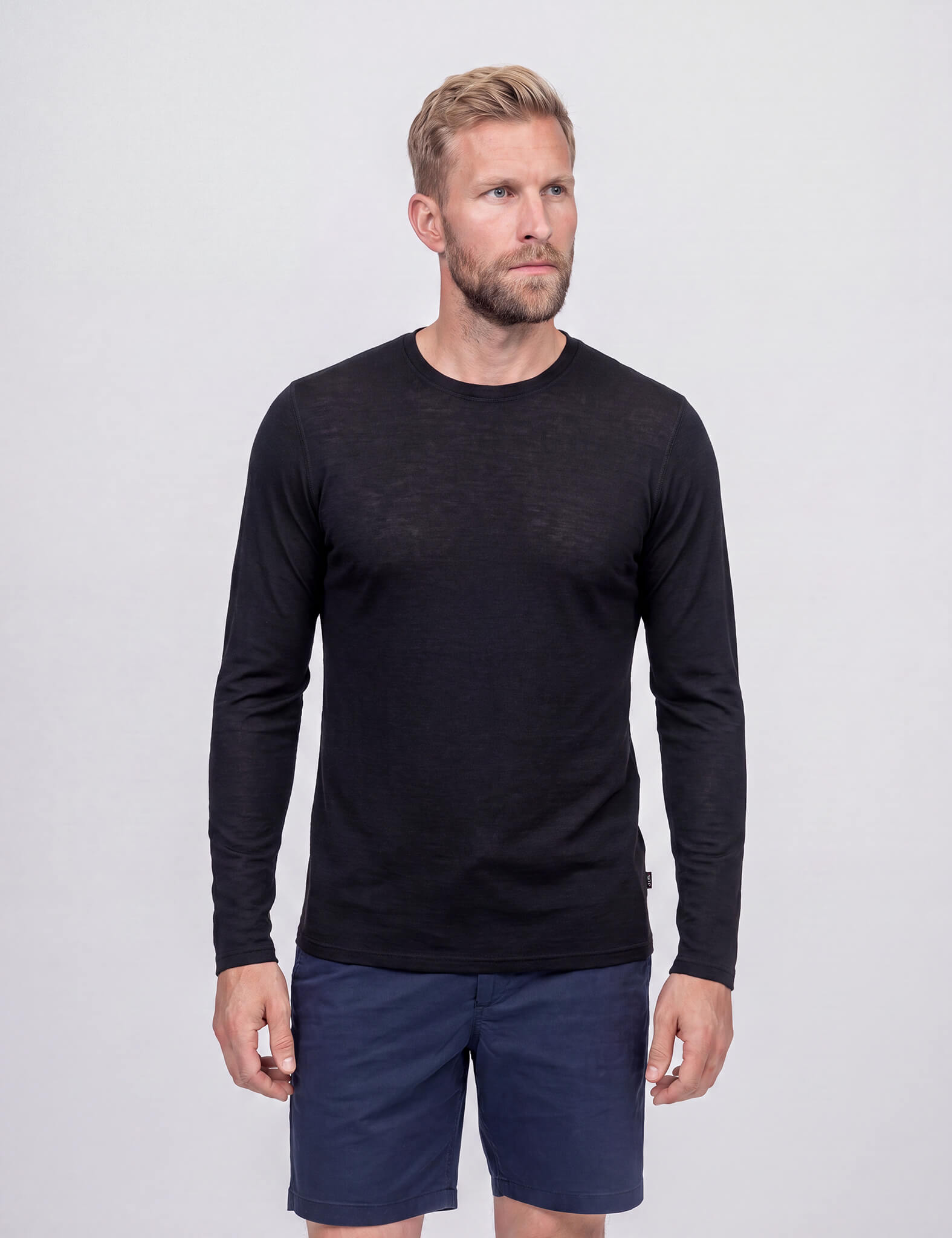 A man with blonde hair and a beard wears the Crew Neck – Classic Merino 160 Men in black along with navy blue shorts, standing against a plain light gray background.