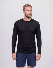 A man with blonde hair and a beard wears the Crew Neck – Classic Merino 160 Men in black along with navy blue shorts, standing against a plain light gray background.