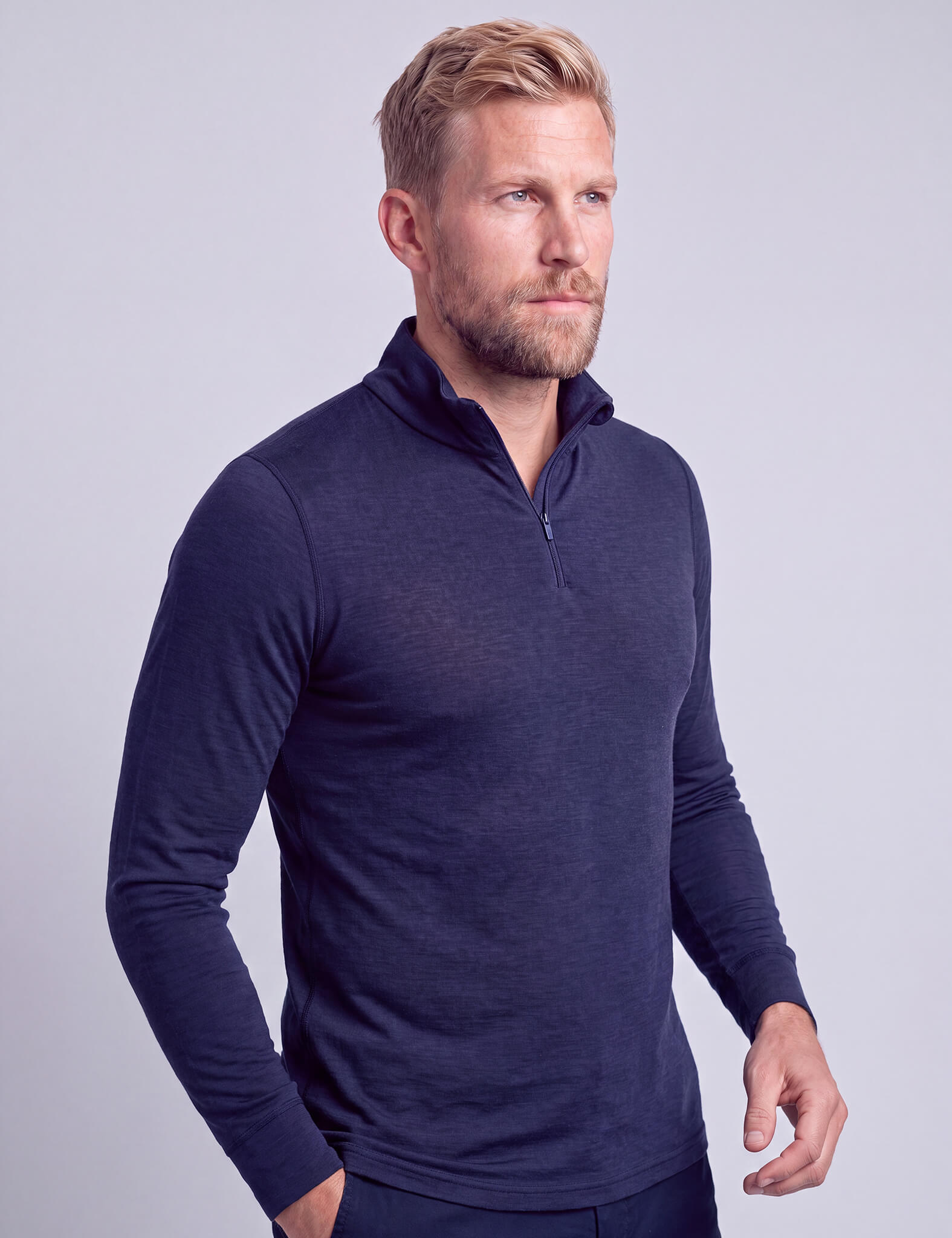 A man with blond hair and a beard is wearing the Half Zip – Classic Merino 160 Men in navy blue along with dark pants, standing against a plain light gray background.