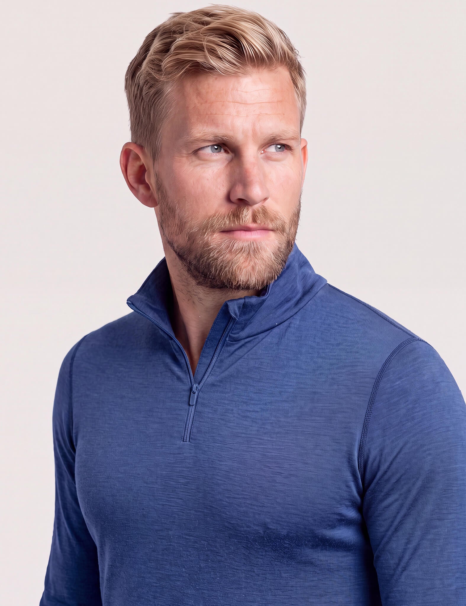 A man with short blonde hair and a beard looks to the side while wearing the Half Zip – Classic Merino 160 Men, a blue long-sleeve shirt designed for natural temperature regulation.