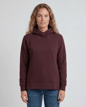 A woman with wavy, light brown hair wears the maroon Hoodie Merino – AirWool 280 Women and blue jeans, standing against a plain light gray background and facing the camera—ideal for cold weather.