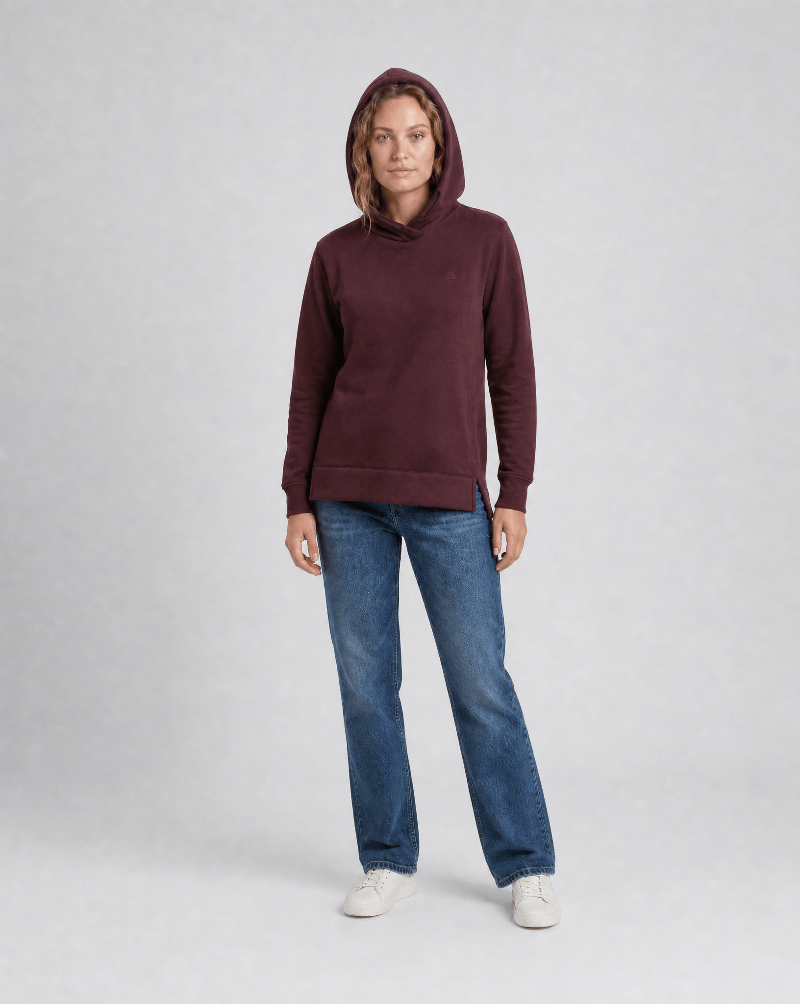 A woman wears the Hoodie Merino – AirWool 280 Women with the hood up, paired with blue jeans and white sneakers against a light gray background—showcasing this stylish, insulating hoodie ideal for cold weather.