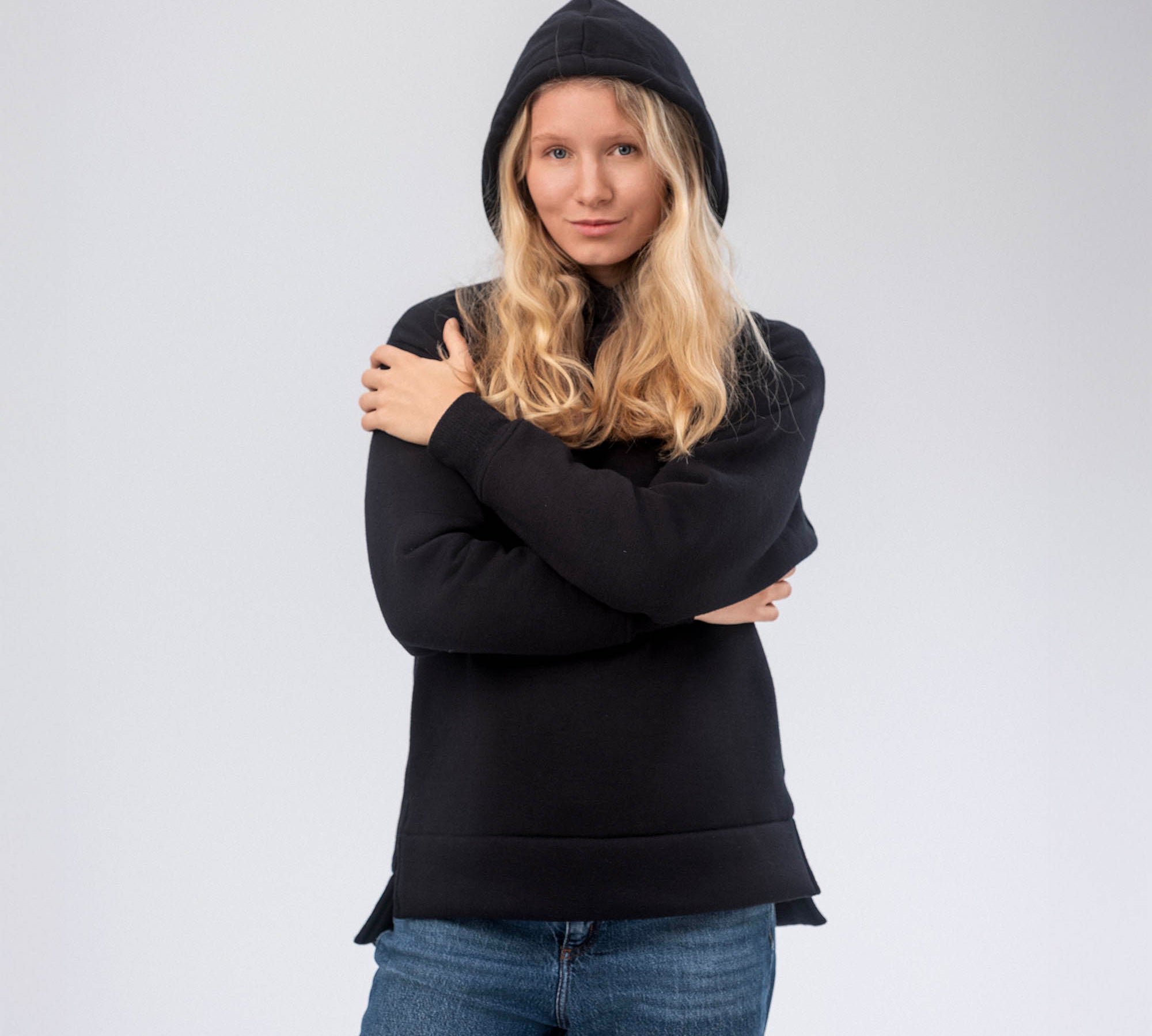 A woman with long blonde hair models the Hoodie Merino – AirWool 280 Women in black, displaying insulating apparel ideal for cool to cold conditions, as she stands with arms crossed against a plain light background.