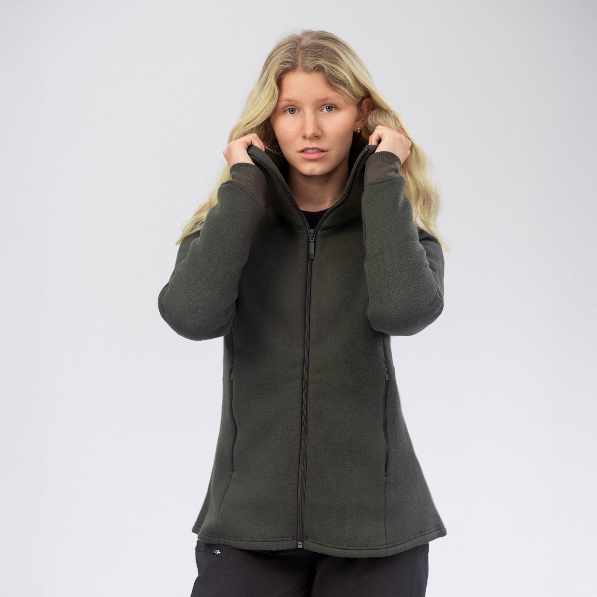 A woman with long blonde hair stands against a plain background, wearing the AirWool 280 Women Hooded Jacket Merino in Forest Night. She holds the high collar with both hands and looks directly at the camera.