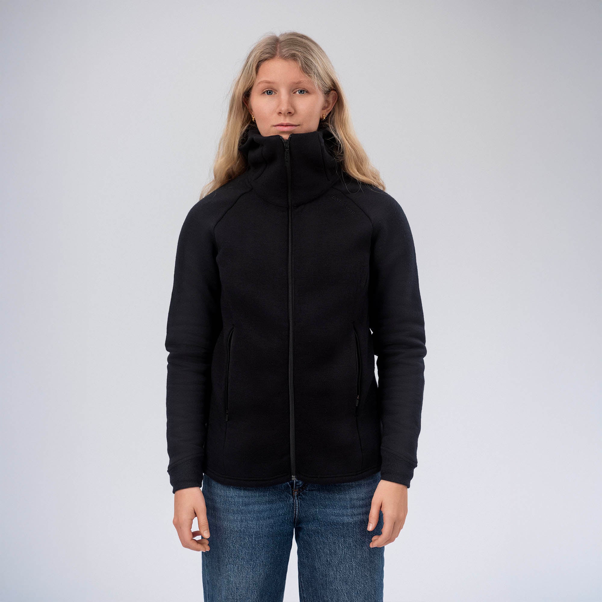 A woman with long blonde hair wearing a black Hooded Jacket Merino – AirWool 280 Women and blue jeans stands facing forward against a plain light gray background.