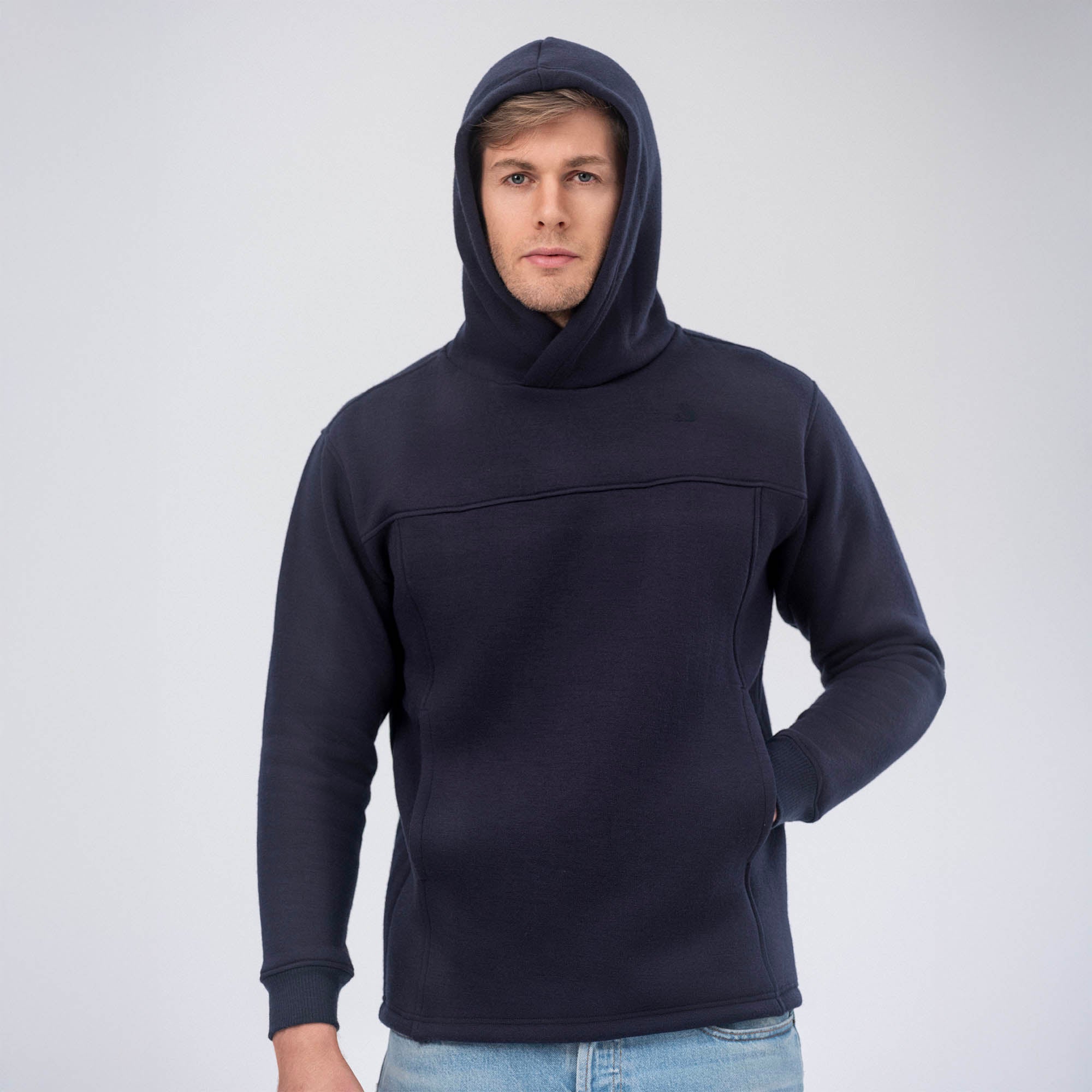 A person stands against a light gray background wearing the Hoodie Merino – AirWool 280 Men in dark navy blue, hood up. One hand is in a pocket while the other arm rests at their side, showcasing this versatile merino wool hoodie.