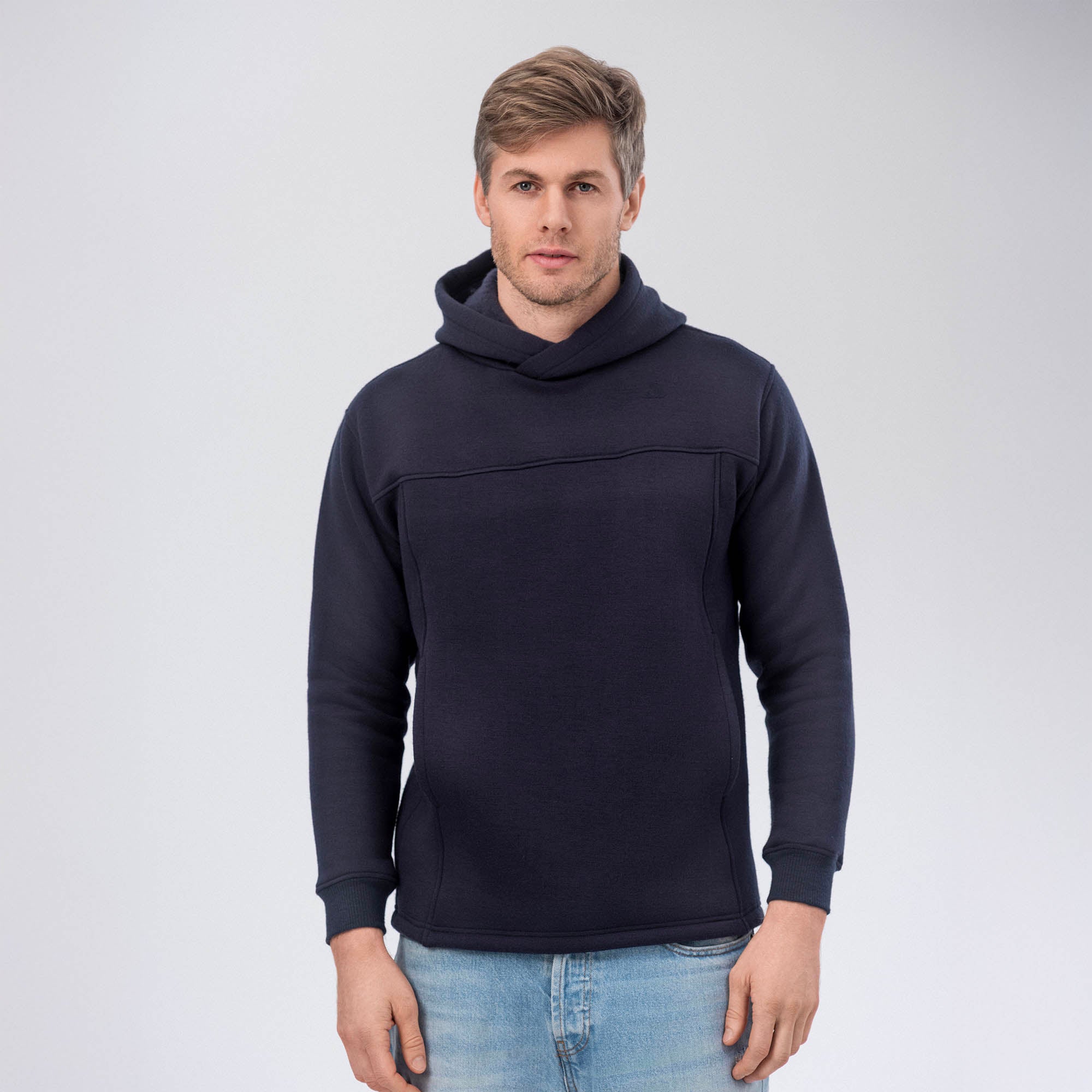 A man with light brown hair, wearing the Hoodie Merino – AirWool 280 Men and light blue jeans, stands facing forward against a plain light gray background.