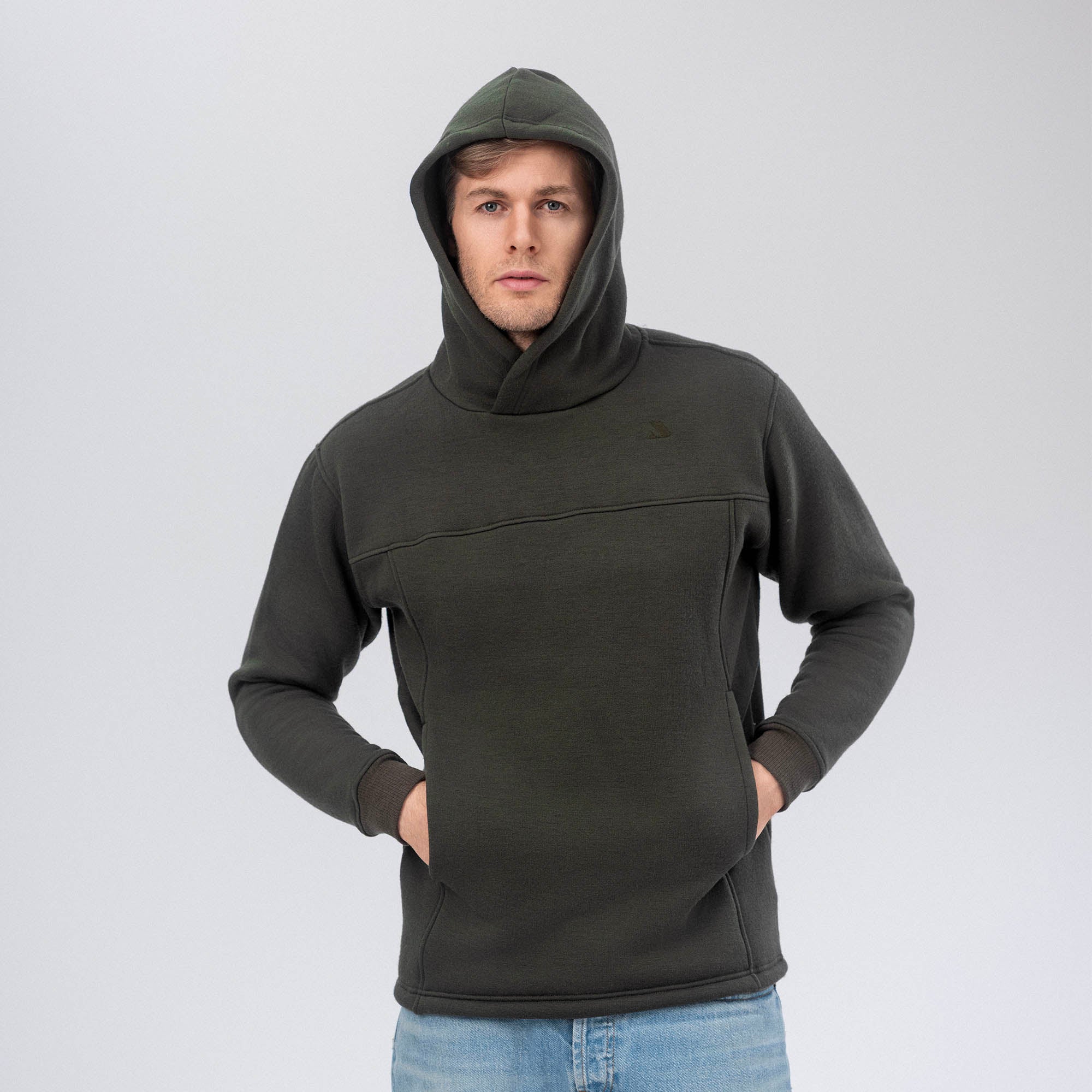 A man stands facing forward against a plain light gray background, wearing the Hoodie Merino – AirWool 280 Men in dark green with his hands in the front pocket.