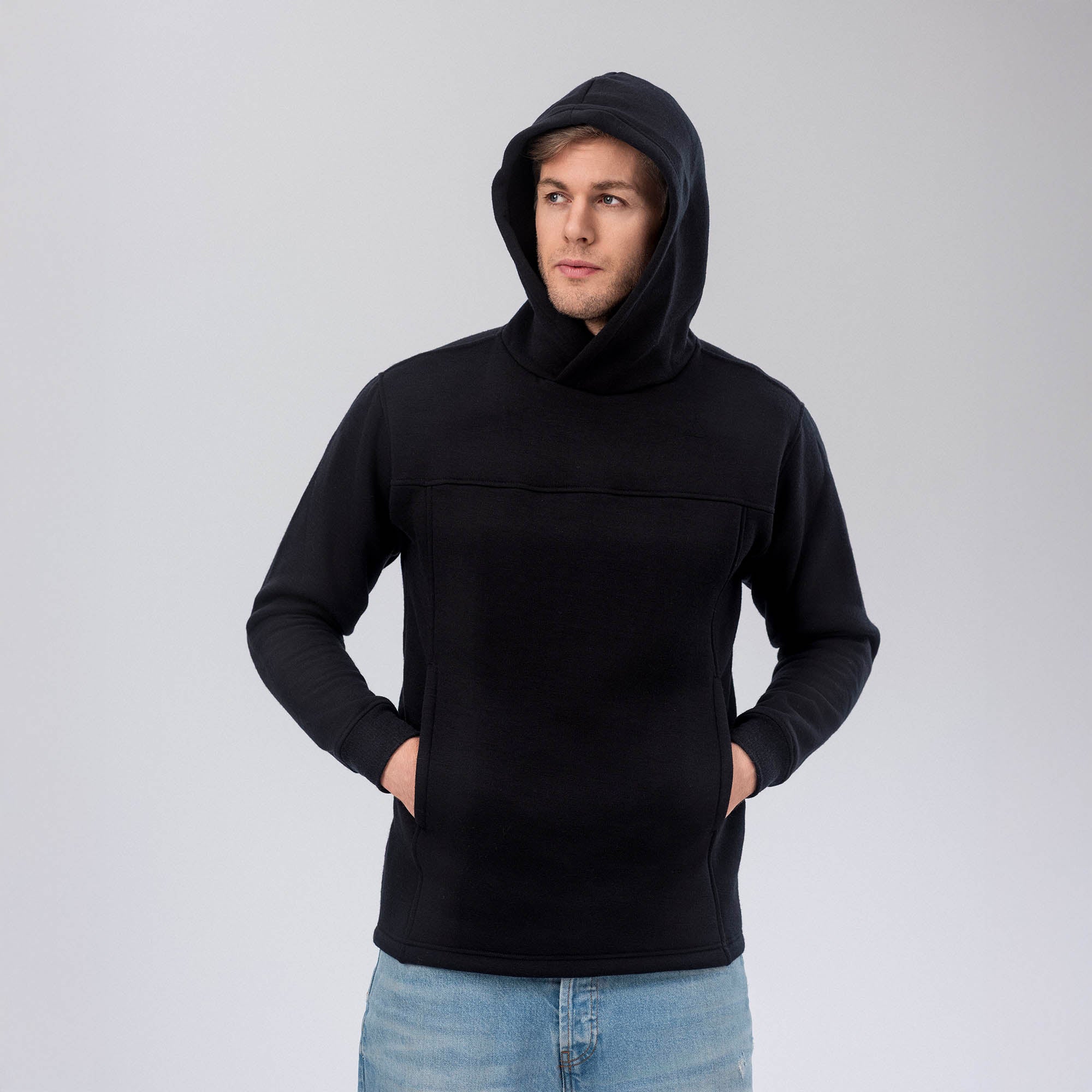 A person stands against a plain, light gray background, wearing the Hoodie Merino – AirWool 280 Men in black with the hood up and hands in the front pocket, looking slightly to the side.
