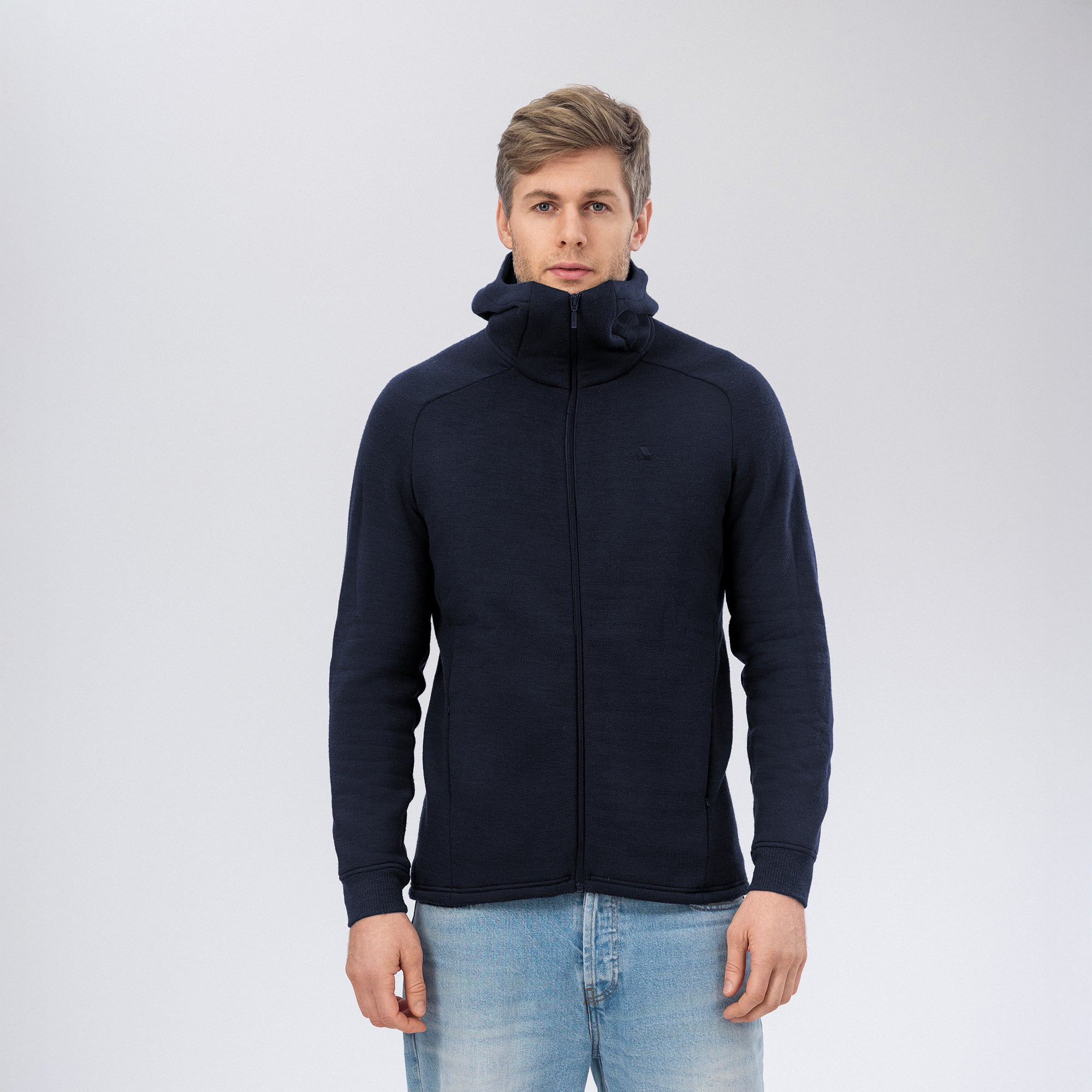 A man with short blond hair faces forward, wearing the Navy Hooded Jacket Merino – AirWool 280 Men and light blue jeans against a plain light background.