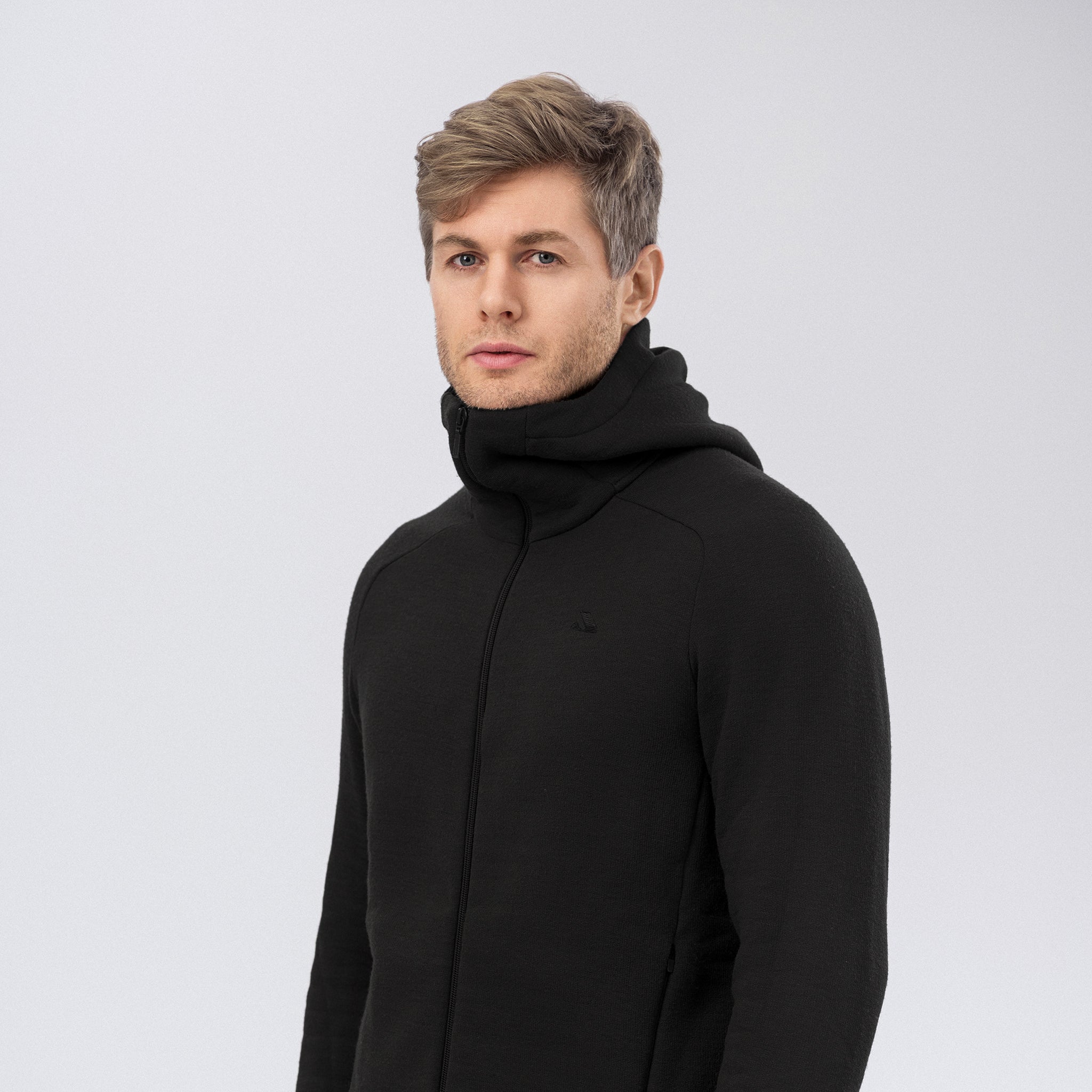 A young man with short, light brown hair wears the black Hooded Jacket Merino – AirWool 280 Men. He stands against a plain, light gray background, looking slightly to the side with a neutral expression.