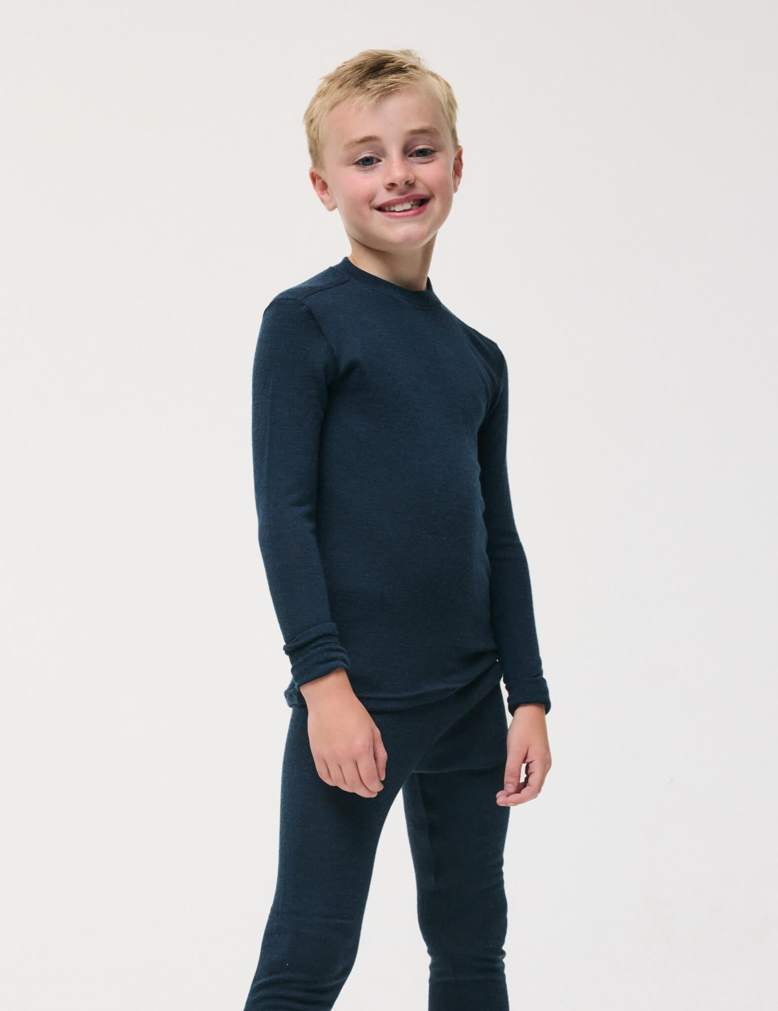 A smiling young boy stands against a plain white background, wearing the Crewneck – Merino Kids Classic Rib in navy, featuring a matching long-sleeve top and pants made from soft ribbed wool jersey.
