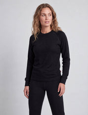 A woman wearing a black Wool Set – Classic Merino 200 Rib Women consisting of a long-sleeved, ribbed merino wool crew-neck top and matching leggings.