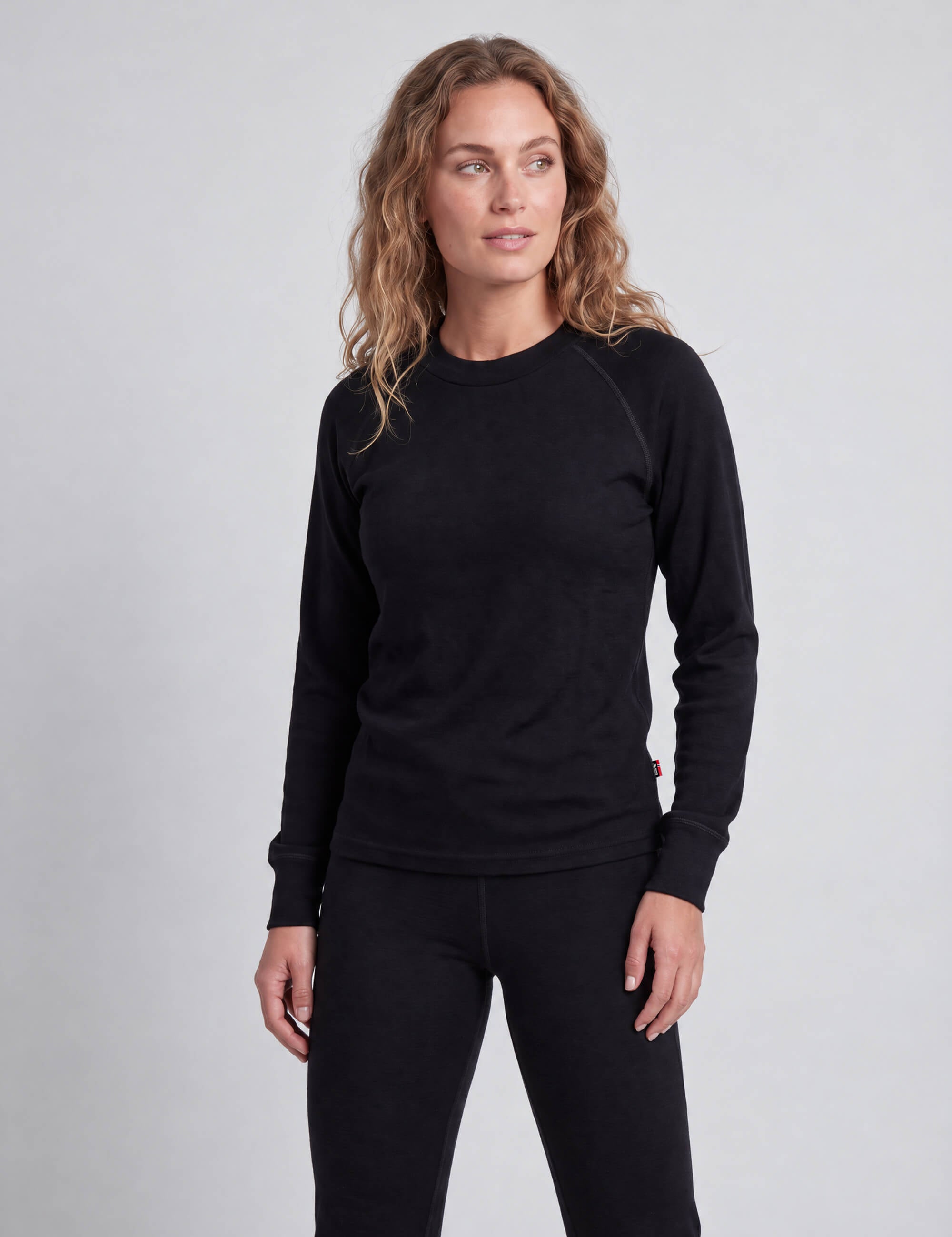 A woman wearing a black Wool Set – Classic Merino 200 Rib Women consisting of a long-sleeved, ribbed merino wool crew-neck top and matching leggings.