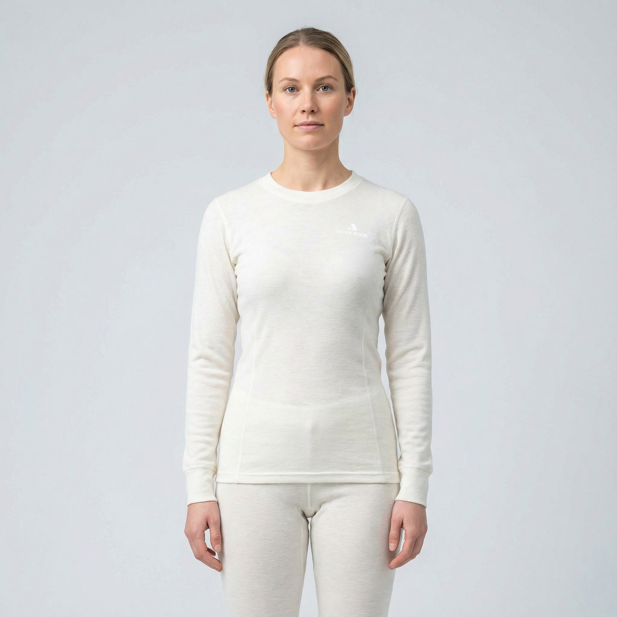 A woman stands facing forward against a plain light gray background, wearing the Jersey – Classic Merino 215 Women in Marshmallow, a long-sleeved thermal top and matching pants that offer warmth and breathability.