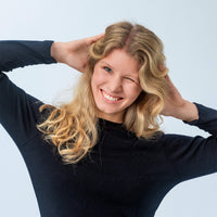 Wearing the Jersey – Classic Merino 215 Women, a smiling woman with long blonde hair poses against a light background, hands behind her head and winking playfully, radiating warmth and effortless breathability.
