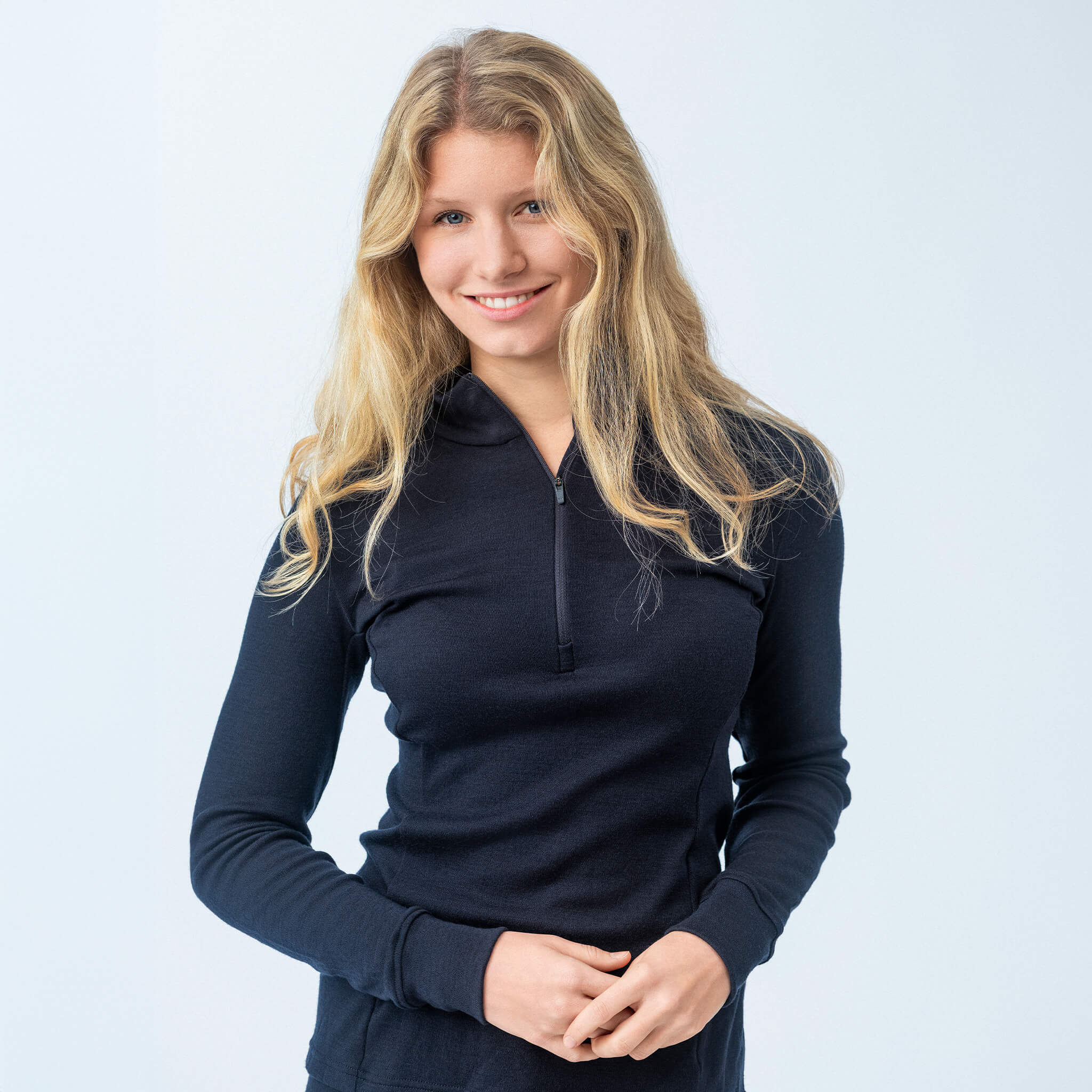 A young woman with long blonde hair wears the Polo Zip – Classic Merino 215 Women in navy, standing against a light background and smiling at the camera with her hands gently clasped in front of her.