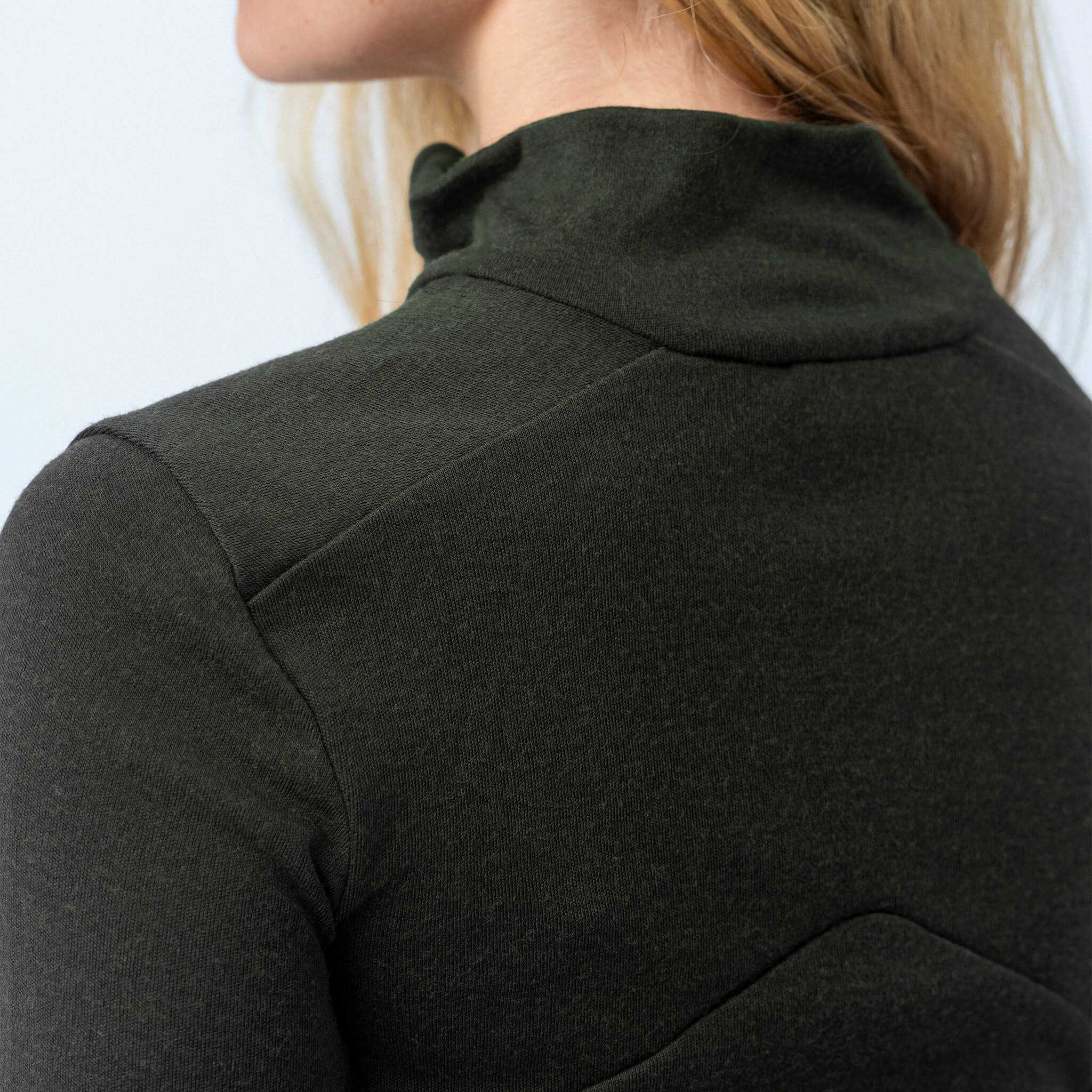 A close-up of a person wearing the Polo Zip – Classic Merino 215 Women in dark green, showing the upper back, shoulder, and neck. Blonde hair stands out against a light-colored background.