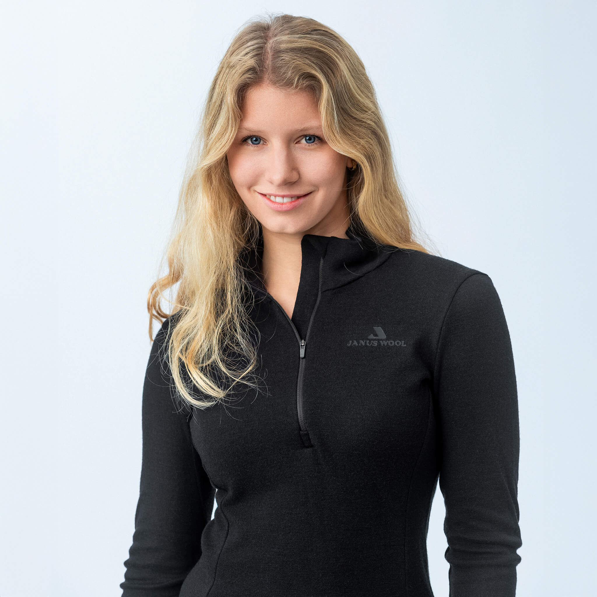 A woman with long blonde hair stands against a plain light background, smiling softly at the camera while wearing the Polo Zip – Classic Merino 215 Women in black.