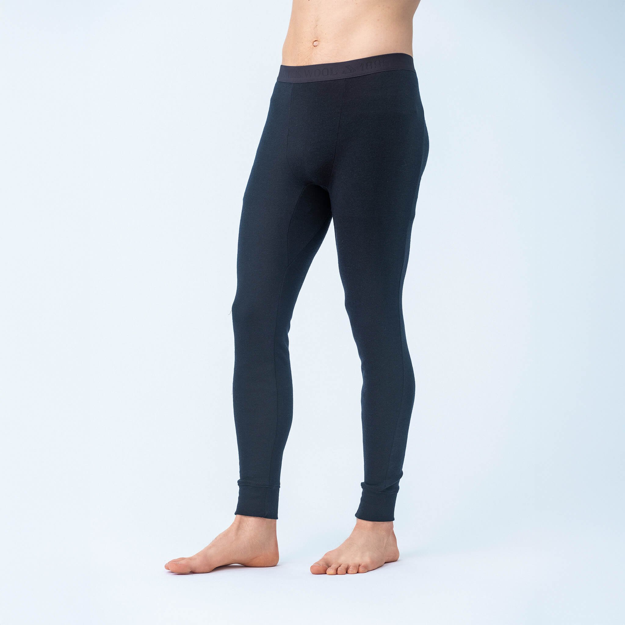 A person wears navy Leggings – Classic Merino 215 Men, standing barefoot against a plain white background. The cropped image focuses on the legs and lower torso, showcasing the leggings’ warmth and breathability.