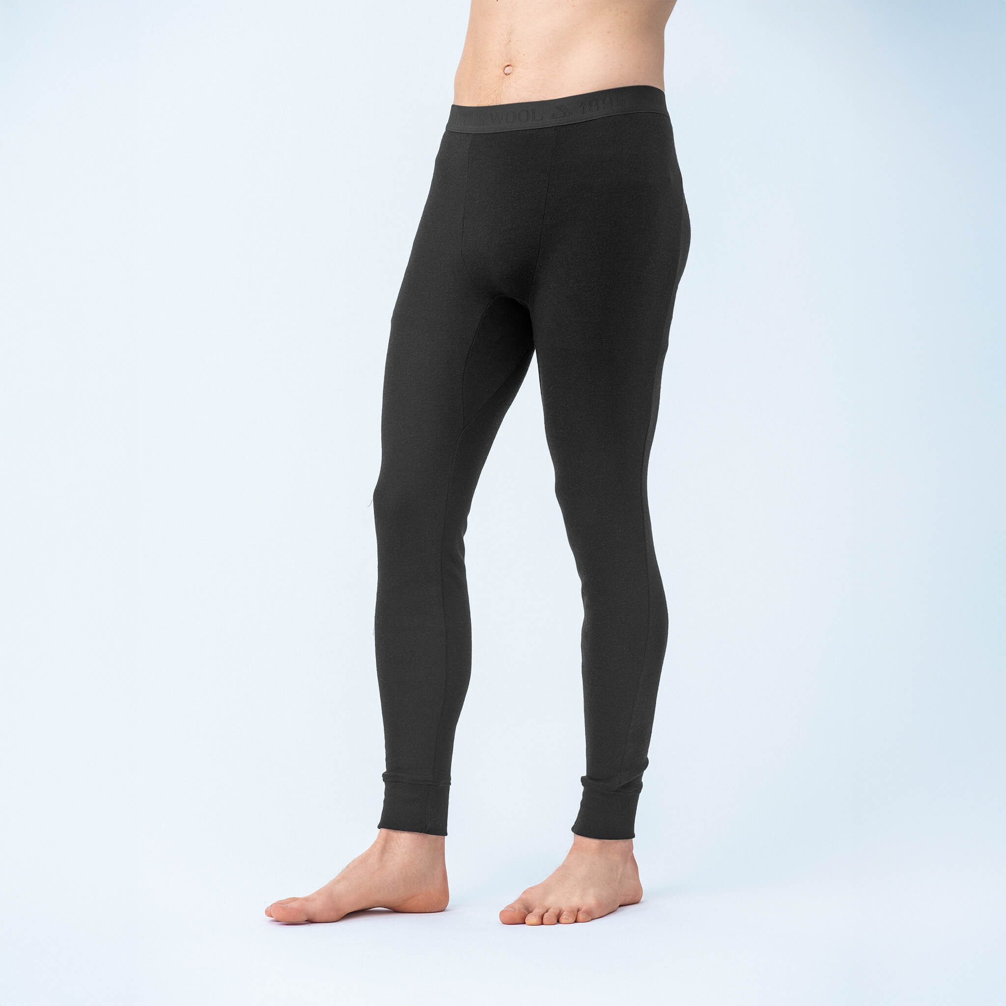 A barefoot man stands against a plain, light background, wearing black Leggings – Classic Merino 215 Men with an elastic waistband and cuffed ankles. The leggings offer warmth and breathability. His upper body is not visible.