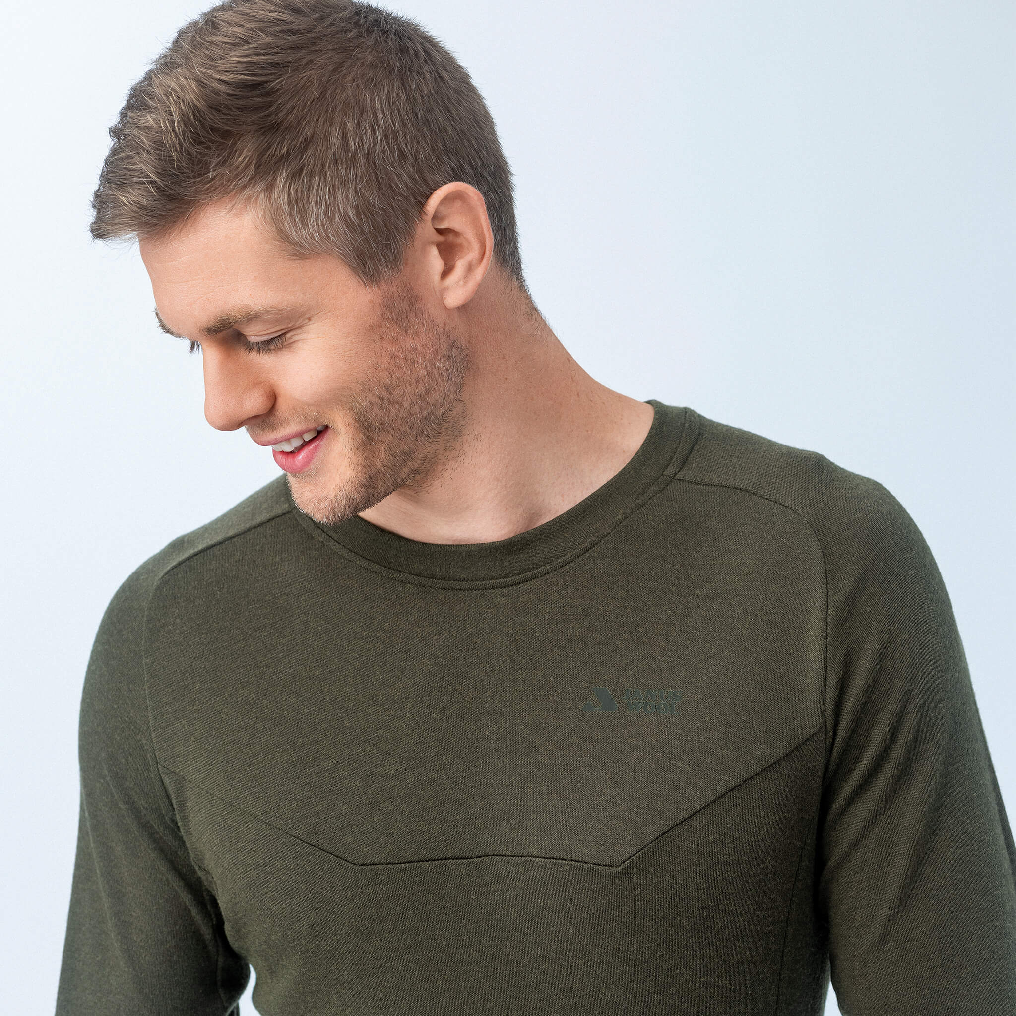 A man with short light brown hair smiles, looking down to his left. He wears the fitted, long-sleeve Jersey – Classic Merino 215 Men in olive green, set against a plain light background.