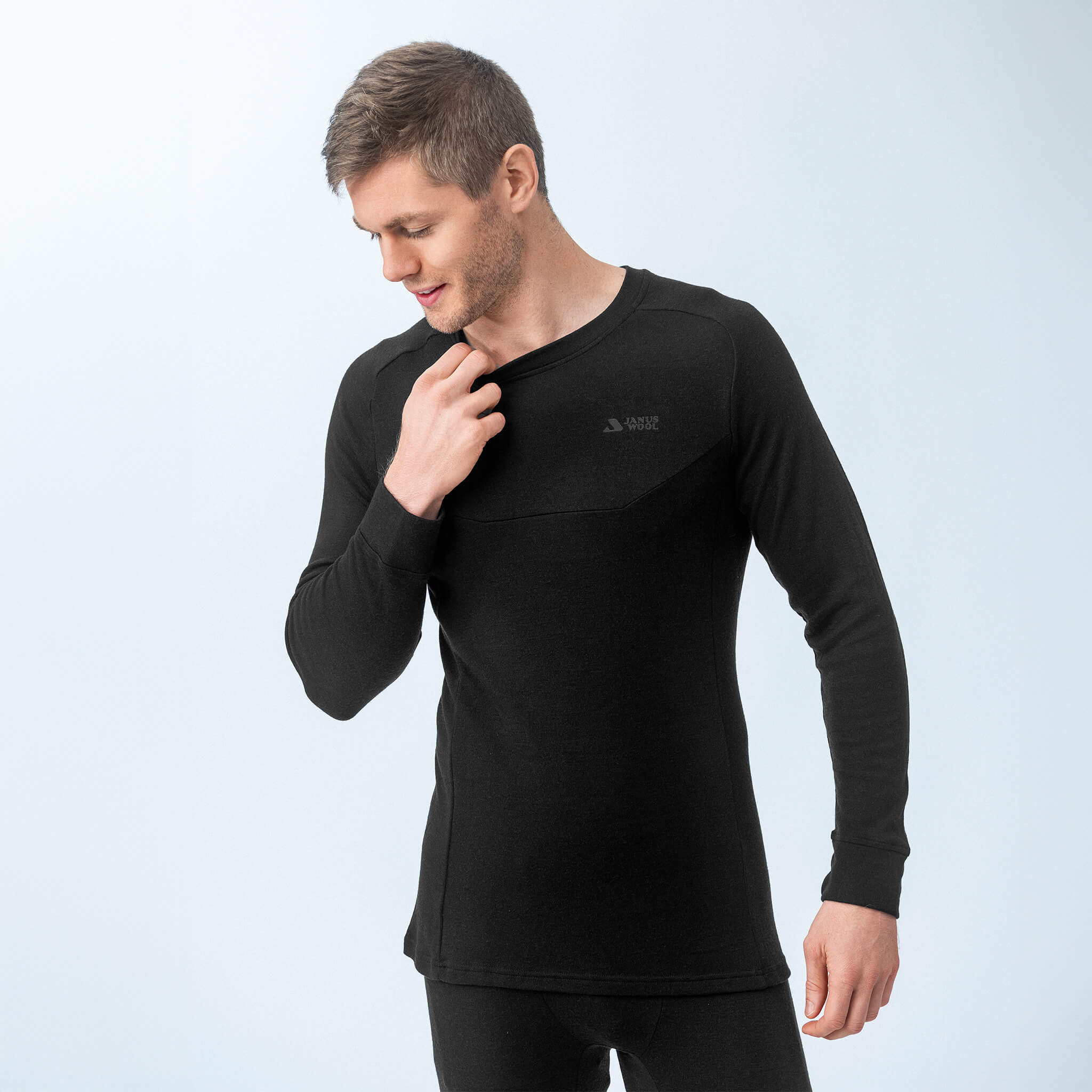 A man wearing the Jersey – Classic Merino 215 Men, a fitted black merino wool long-sleeve top with subtle chest branding, stands against a light background, looking down and adjusting the collar with one hand.