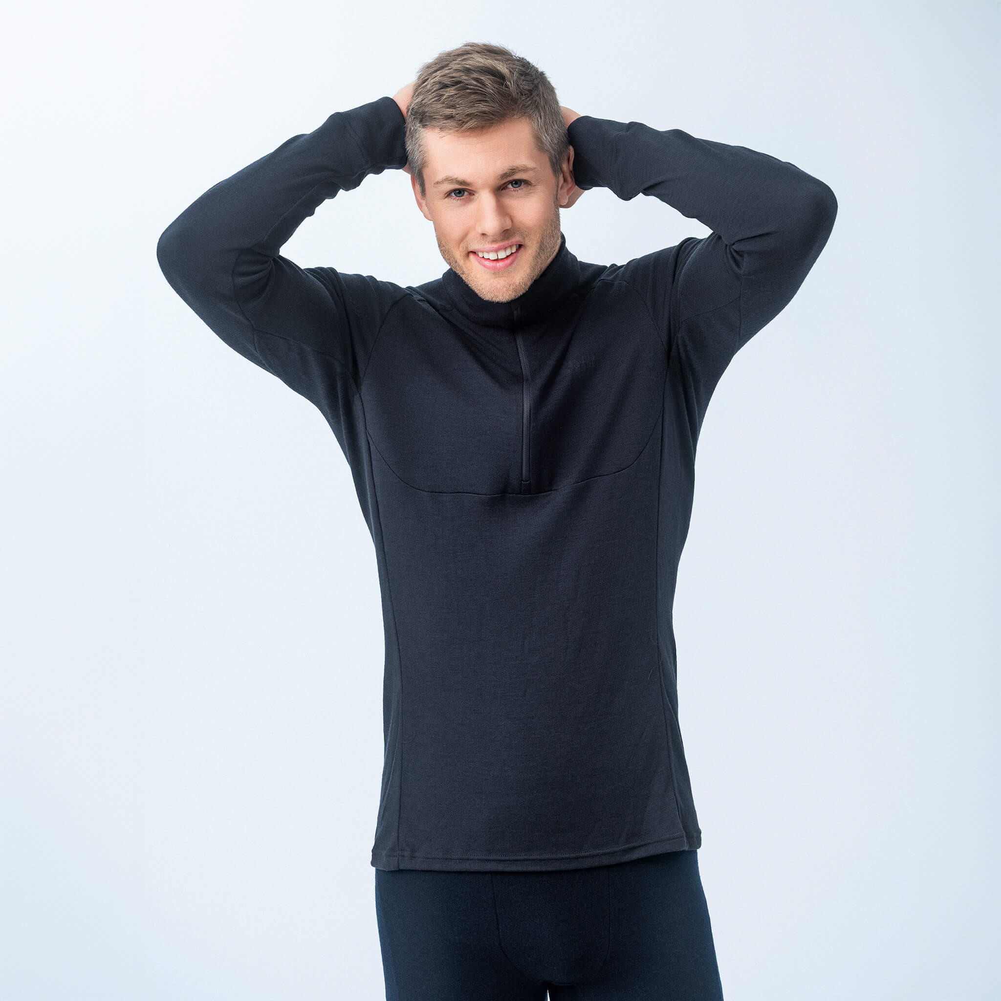 A smiling man in a navy Polo Zip – Classic Merino 215 Men sweater poses with both hands behind his head against a plain light background.