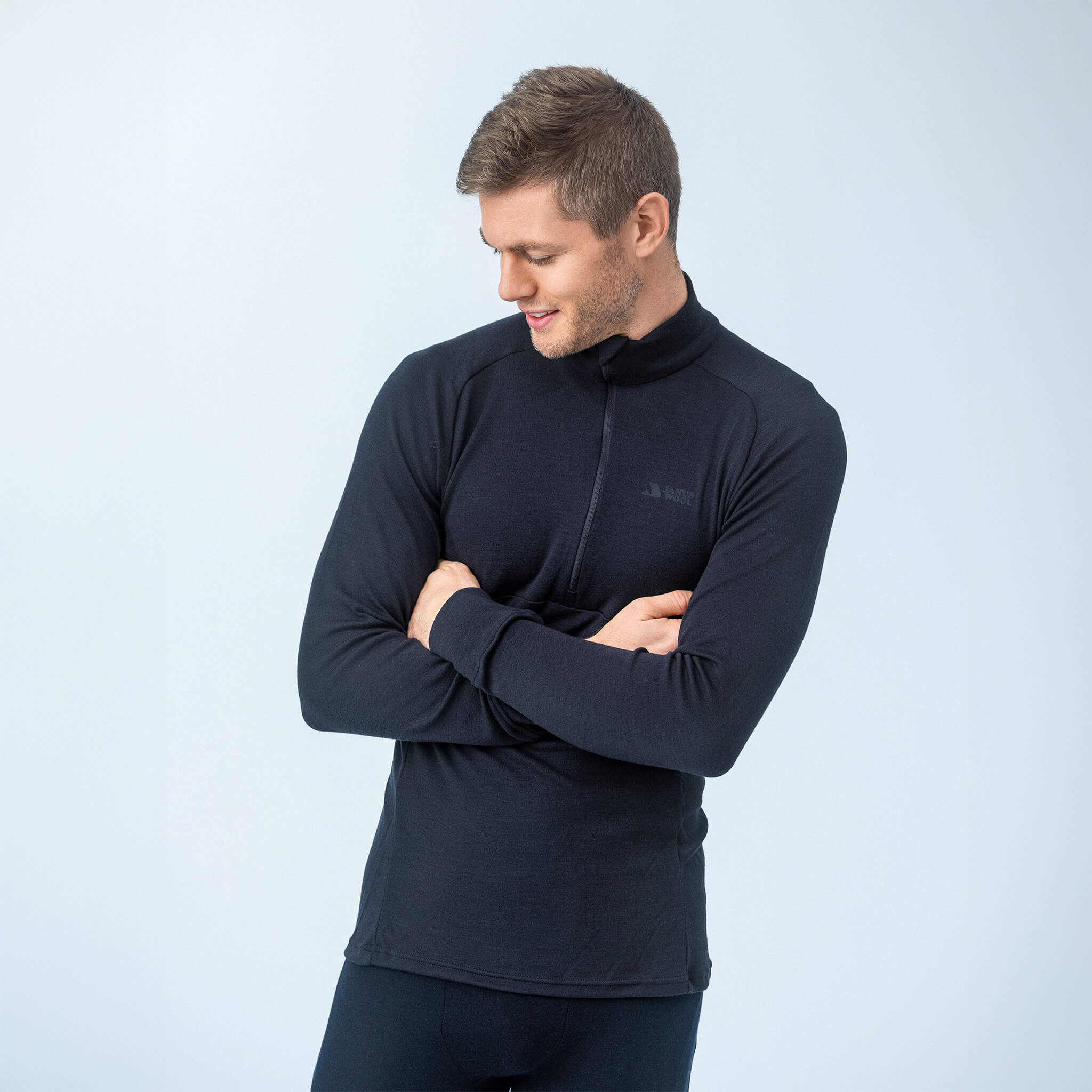 A man stands with arms crossed, looking down to his left and smiling. He is wearing the Polo Zip – Classic Merino 215 Men in black with dark pants against a plain light background.