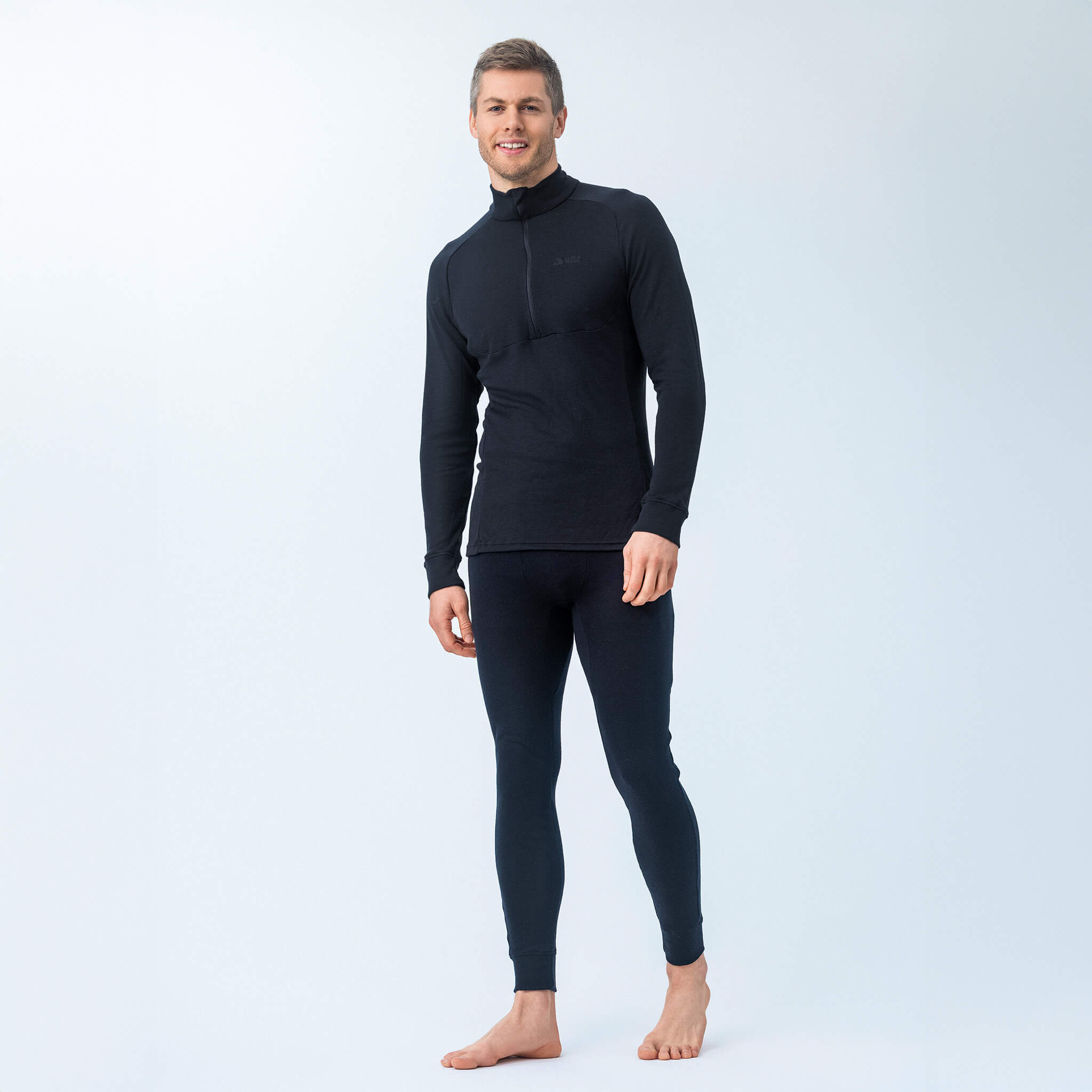 A man stands barefoot against a light background, smiling slightly to the side, wearing the Polo Zip – Classic Merino 215 Men in black with matching black leggings.
