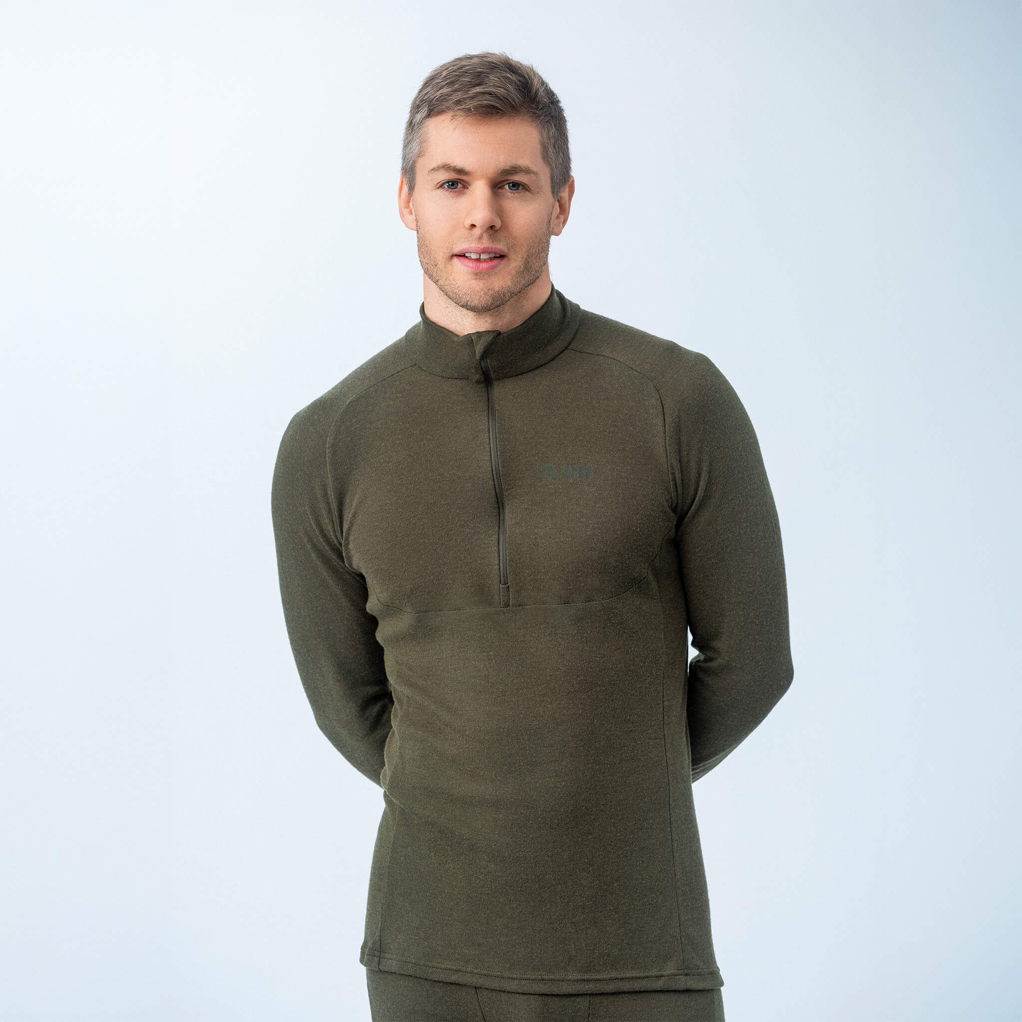 A young man with short hair wears the Polo Zip – Classic Merino 215 Men in Forest Night, standing against a plain light background with his hands behind his back.