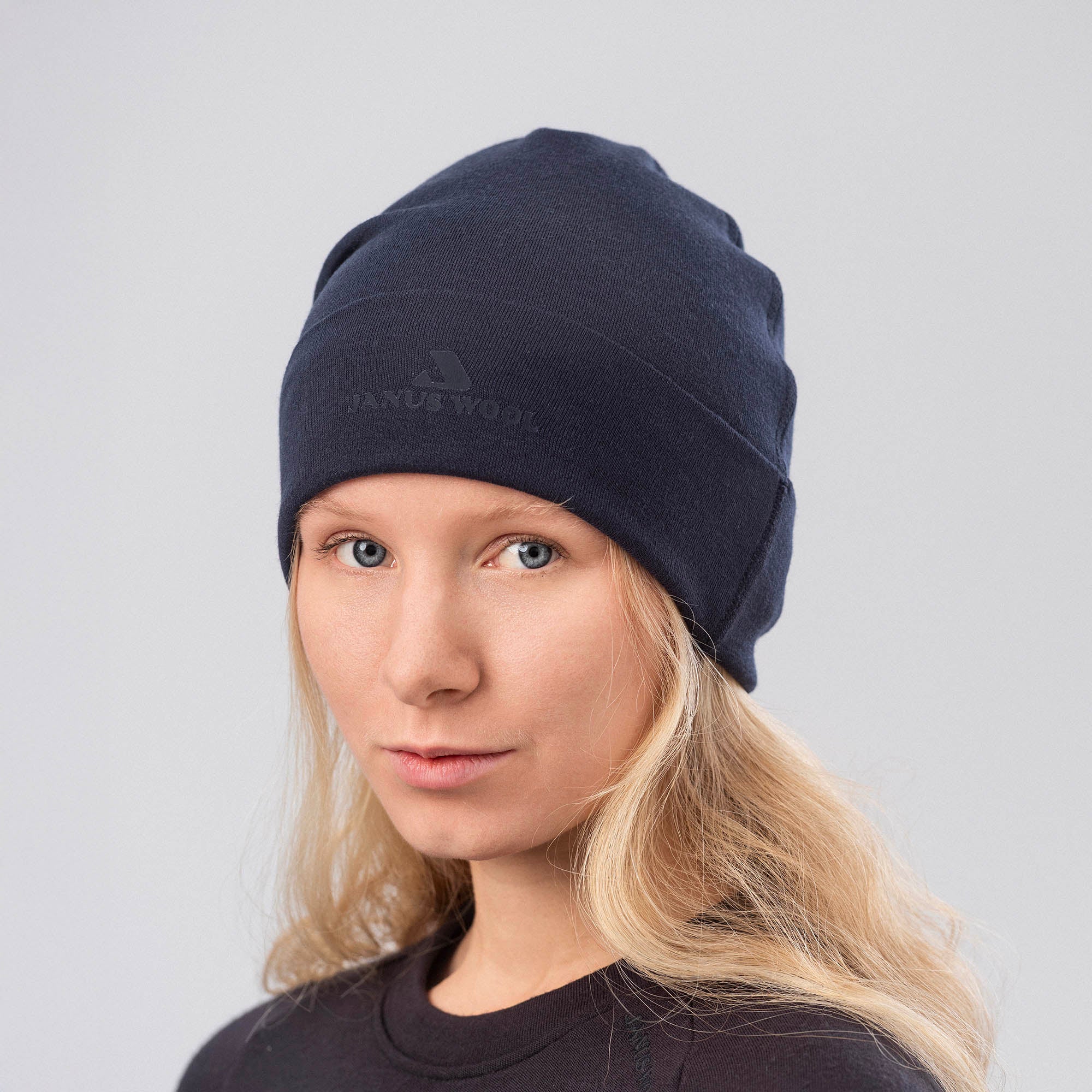 A woman with long blonde hair models the Beanie – Classic Merino 215 in dark blue and a matching shirt, looking at the camera against a plain light gray background, highlighting the beanie's warmth and breathability.