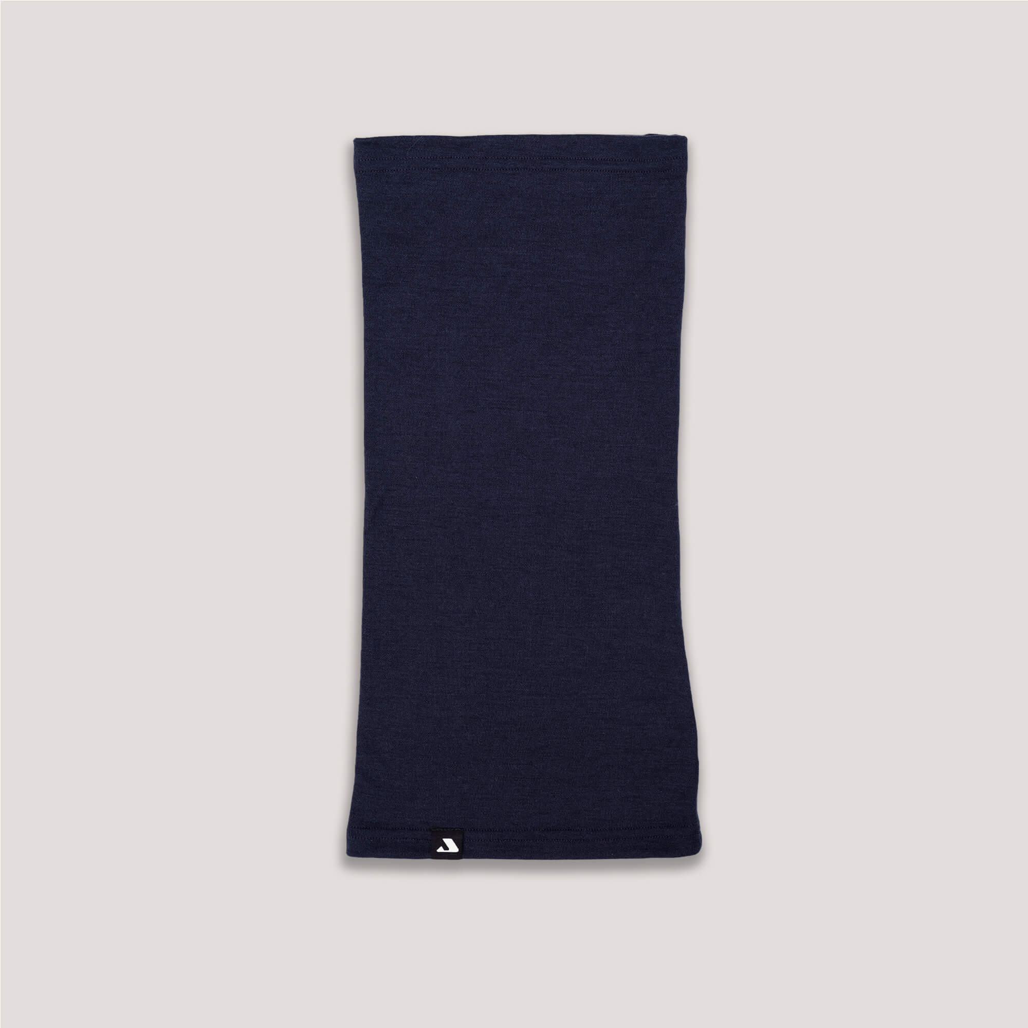The Neck Warmer – Classic Merino 215 in navy is shown laid flat on a light gray surface, featuring a small white logo tag near the bottom edge. Its breathable merino wool design offers both comfort and warmth.
