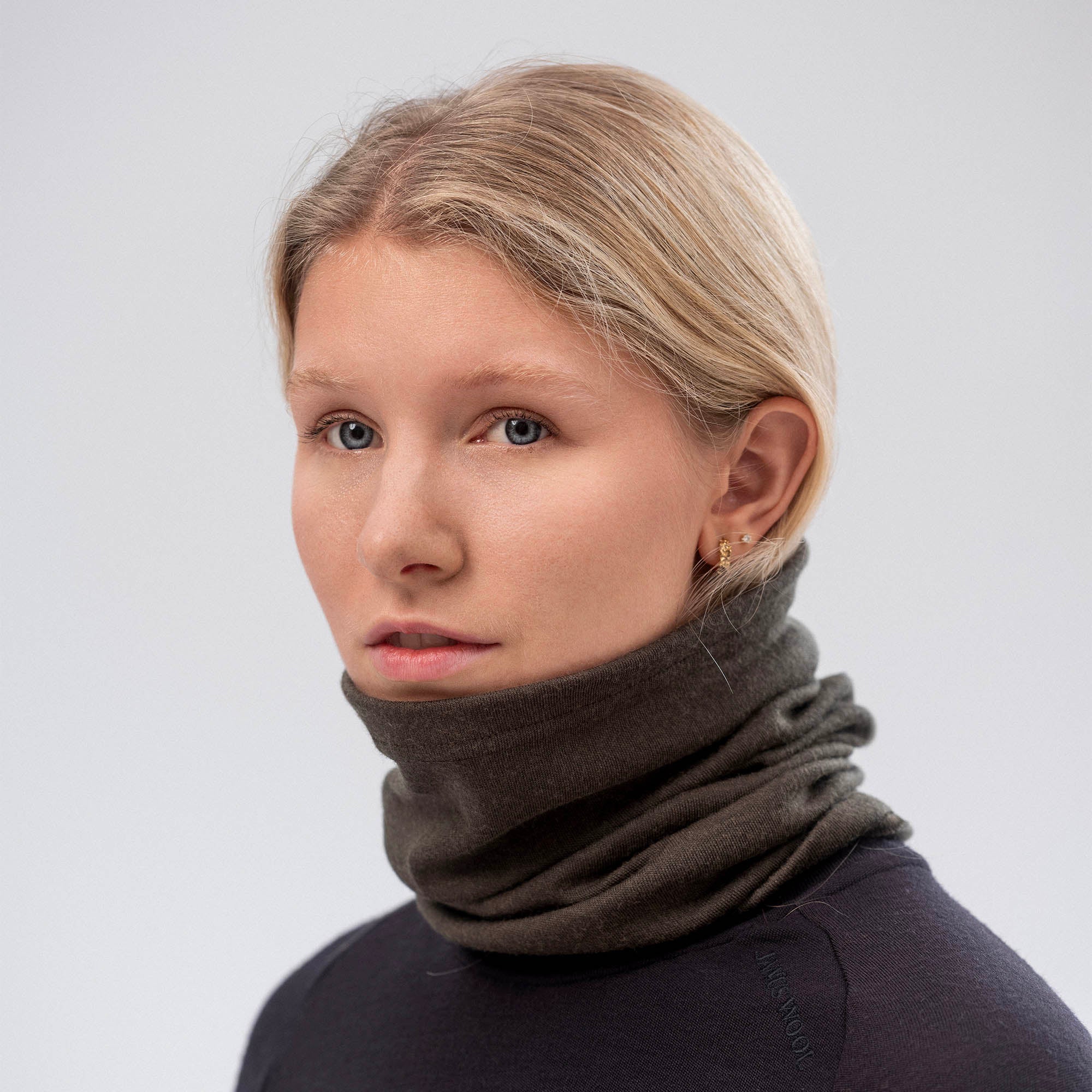 A young blonde woman wears the Neck Warmer – Classic Merino 215, a breathable dark merino wool accessory, paired with a black top as she looks to the side against a plain light background.