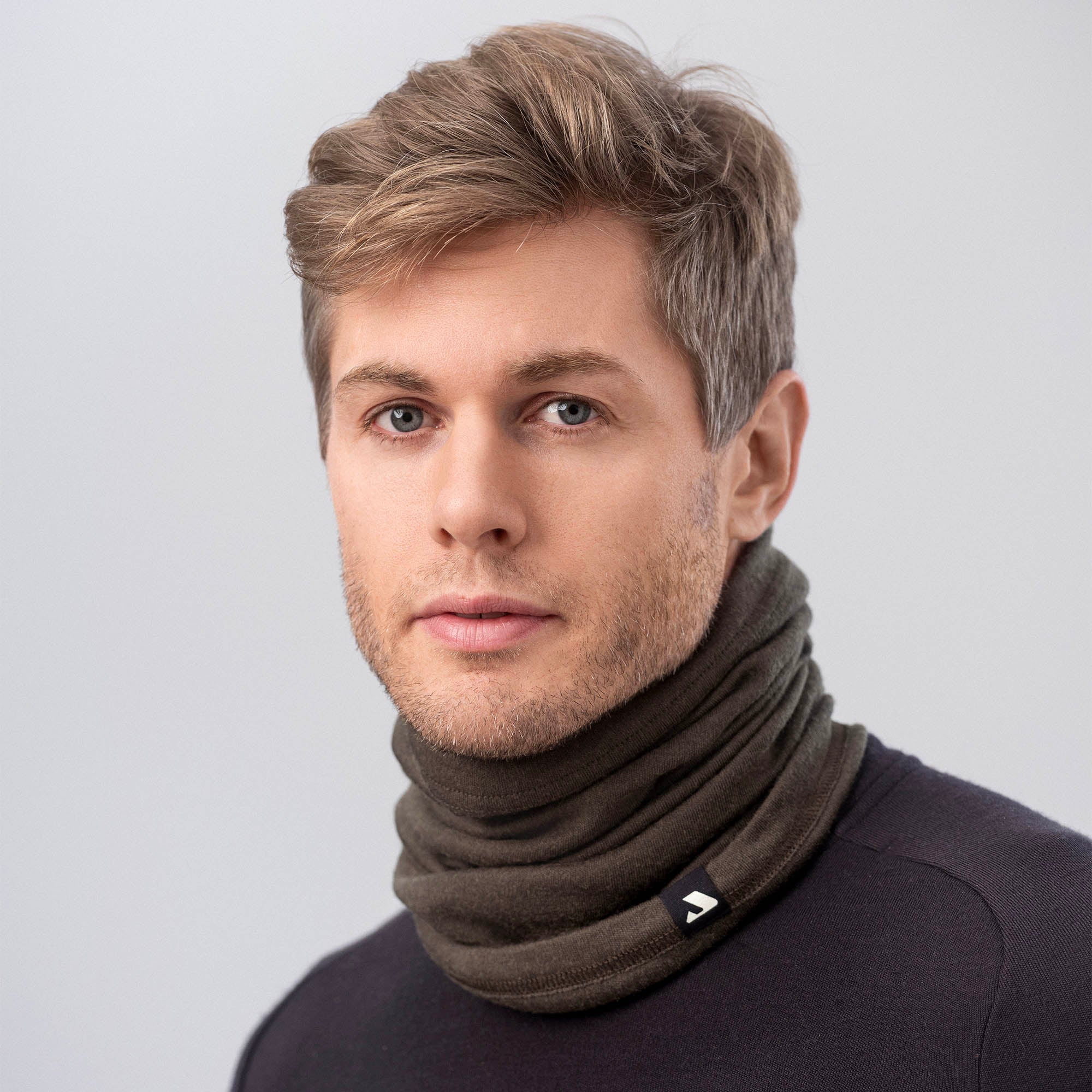 A young man with light brown hair wears the Neck Warmer – Classic Merino 215 and a black shirt, looking directly at the camera against a plain light gray background.