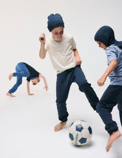 Three children in casual clothes and hats play with a soccer ball on a white background. Two, dressed in Merino Kids AirWool Sweatpants, kick the ball barefoot, while one does a cartwheel in the background.