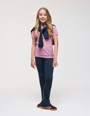A young person with long blonde hair stands smiling against a plain white background, wearing a pink t-shirt, warm and soft Flarepants – Merino Kids AirWool, maroon shoes, and a navy sweater draped over their shoulders.