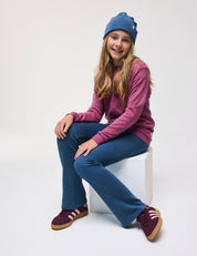 A smiling girl sits on a white cube against a plain background, wearing a blue beanie, a pink long-sleeve shirt, warm and soft Flarepants – Merino Kids AirWool, and maroon sneakers with pink stripes.