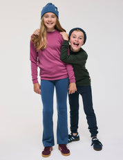 Two smiling children pose together. The girl wears a blue hat, pink sweater, and Flarepants – Merino Kids AirWool, which are warm, soft, and comfortable. The boy, in a black hat and a green sweater made with brushed AirWool paired with dark pants, stands slightly behind her with his arm around her shoulders.