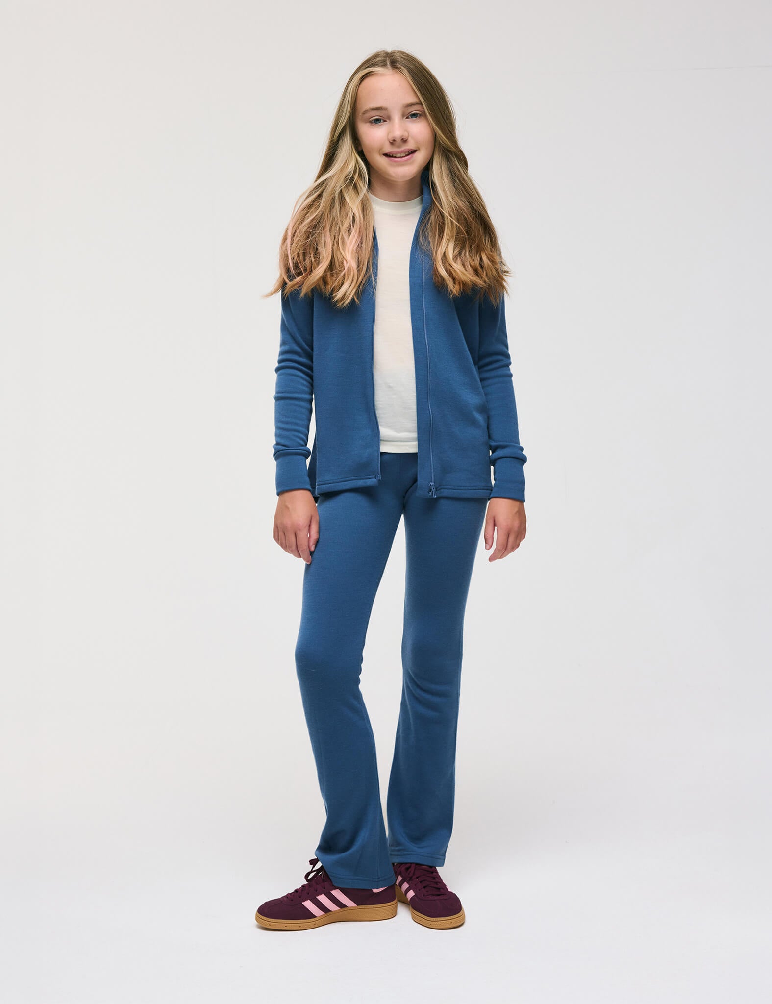 A girl stands against a plain white background, smiling softly as her long, light brown hair frames her face. She wears Flarepants – Merino Kids AirWool, paired with a blue zip-up jacket and a white shirt. For footwear, she chooses burgundy Adidas sneakers. The outfit is made from warm, soft, comfortable materials for everyday wear.