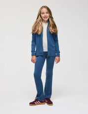 A girl stands against a plain white background, smiling softly as her long, light brown hair frames her face. She wears Flarepants – Merino Kids AirWool, paired with a blue zip-up jacket and a white shirt. For footwear, she chooses burgundy Adidas sneakers. The outfit is made from warm, soft, comfortable materials for everyday wear.