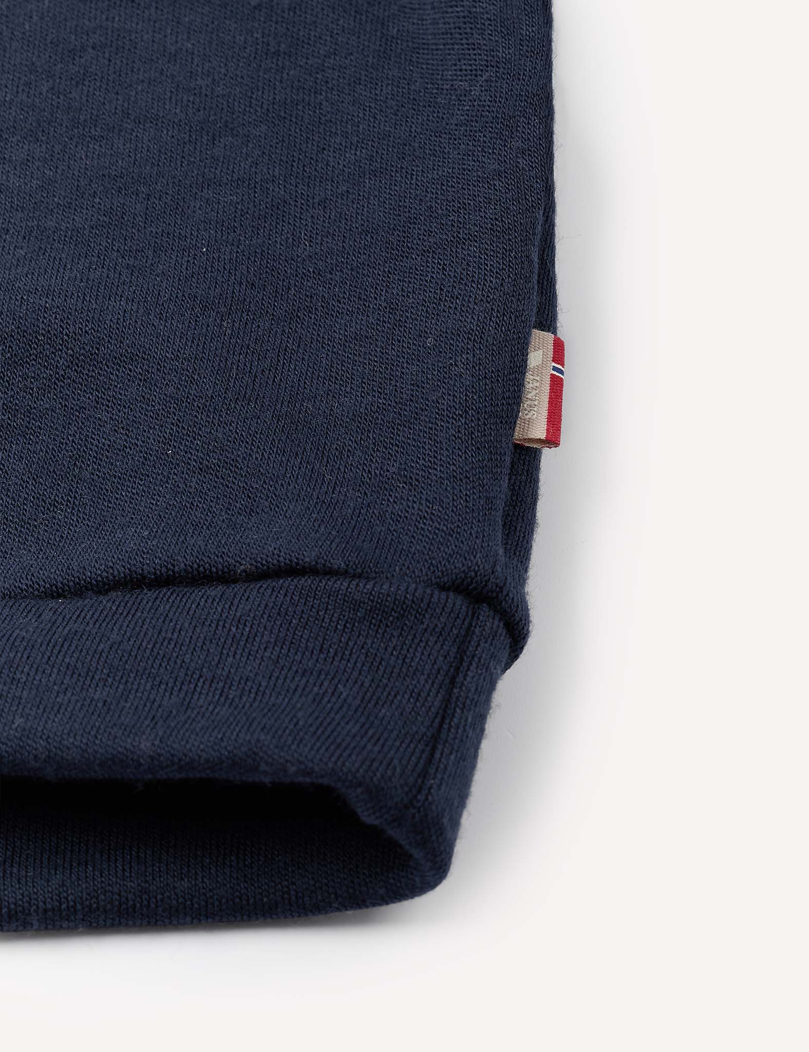 Close-up of a navy blue cuff from the Pullover – Merino Baby AirWool, crafted from soft brushed AirWool fabric, with a small tag featuring a red, white, and blue flag detail stitched into the seam. The background is plain and light-colored.