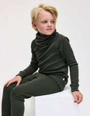 A young child with blonde hair sits on a white block, wearing the Pullover – Merino Kids AirWool in dark green with a loose neck and matching pants, looking off to the side against a plain white background.