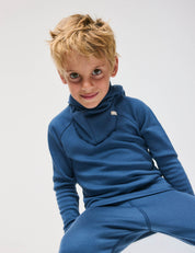A young boy with tousled blond hair wears the Ensign Blue Pullover – Merino Kids AirWool sweater and matching pants, sitting and smiling at the camera against a plain light background.