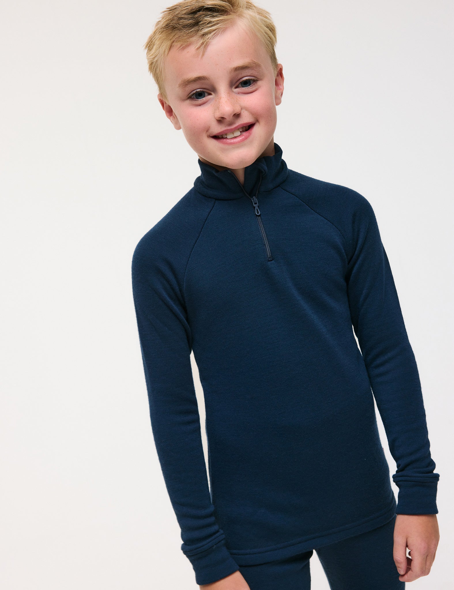 A smiling child with short blond hair wears the Half Zip – Merino Kids AirWool shirt in dark navy blue, paired with matching pants, standing against a plain light background.