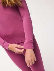A child wearing the mauve Half Zip – Merino Kids AirWool set, featuring a matching long-sleeve top and leggings crafted from brushed AirWool, adjusts the sleeve with one hand against a plain light background.