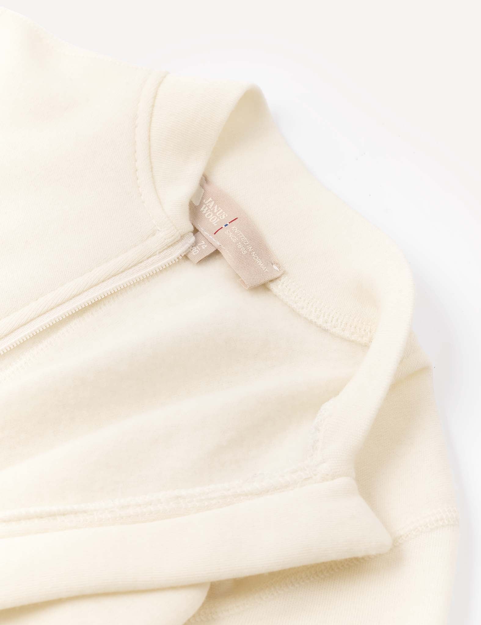 A close-up of the Onesie – Merino Baby AirWool showcases part of the collar, an exposed zipper, and a beige fabric tag with text, all set against a light background. The onesie features fold-over cuffs for added comfort.
