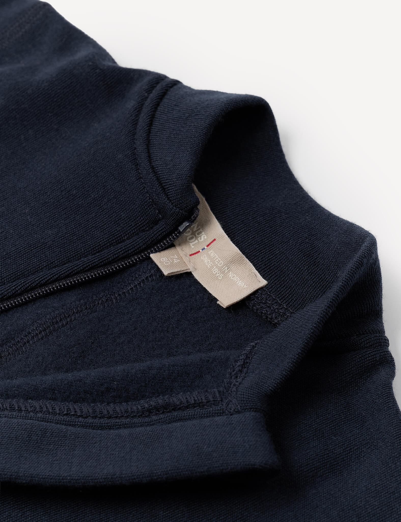 Close-up of the Onesie – Merino Baby AirWool in navy blue, showcasing fold-over cuffs, a visible beige fabric tag, and a short zipper near the collar, laid on a white background.