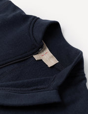 Close-up of the Onesie – Merino Baby AirWool in navy blue, showcasing fold-over cuffs, a visible beige fabric tag, and a short zipper near the collar, laid on a white background.